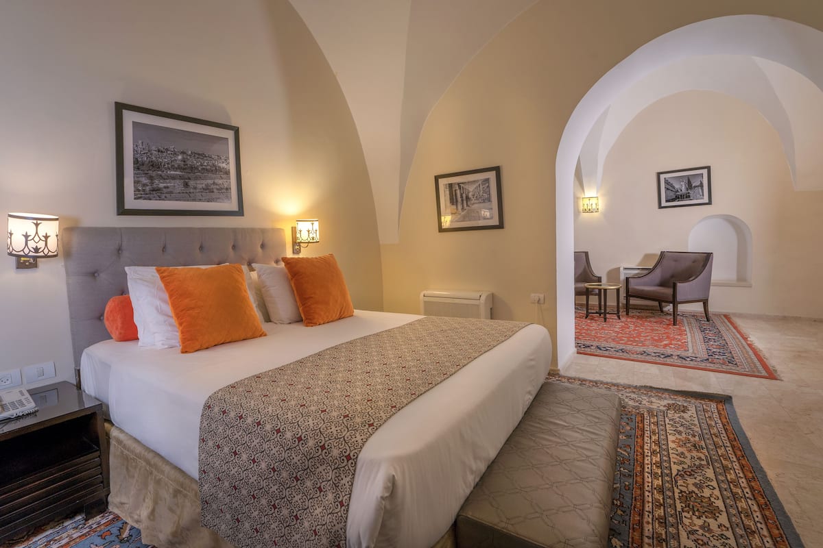 A hotel room with a large bed, two bedside tables, a nightstand, a desk, a chair, and an arched doorway leading to another room.