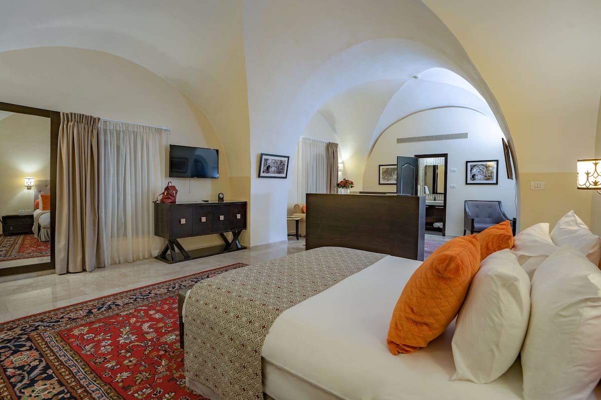 A hotel room with a large bed, a desk, a chair, a TV, and a bathroom visible through an arched doorway.
