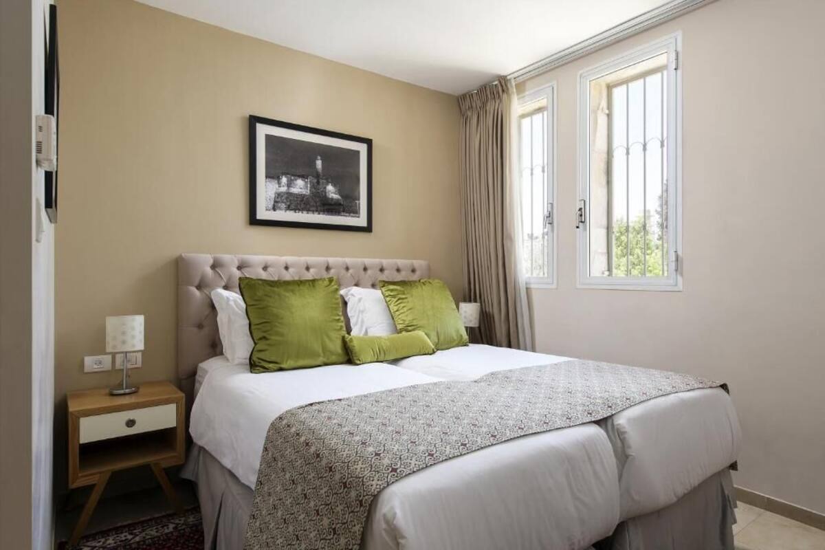 A bedroom with a tufted headboard, a bed with white and green pillows, a nightstand with a lamp, a framed picture on the wall, and a window with curtains.