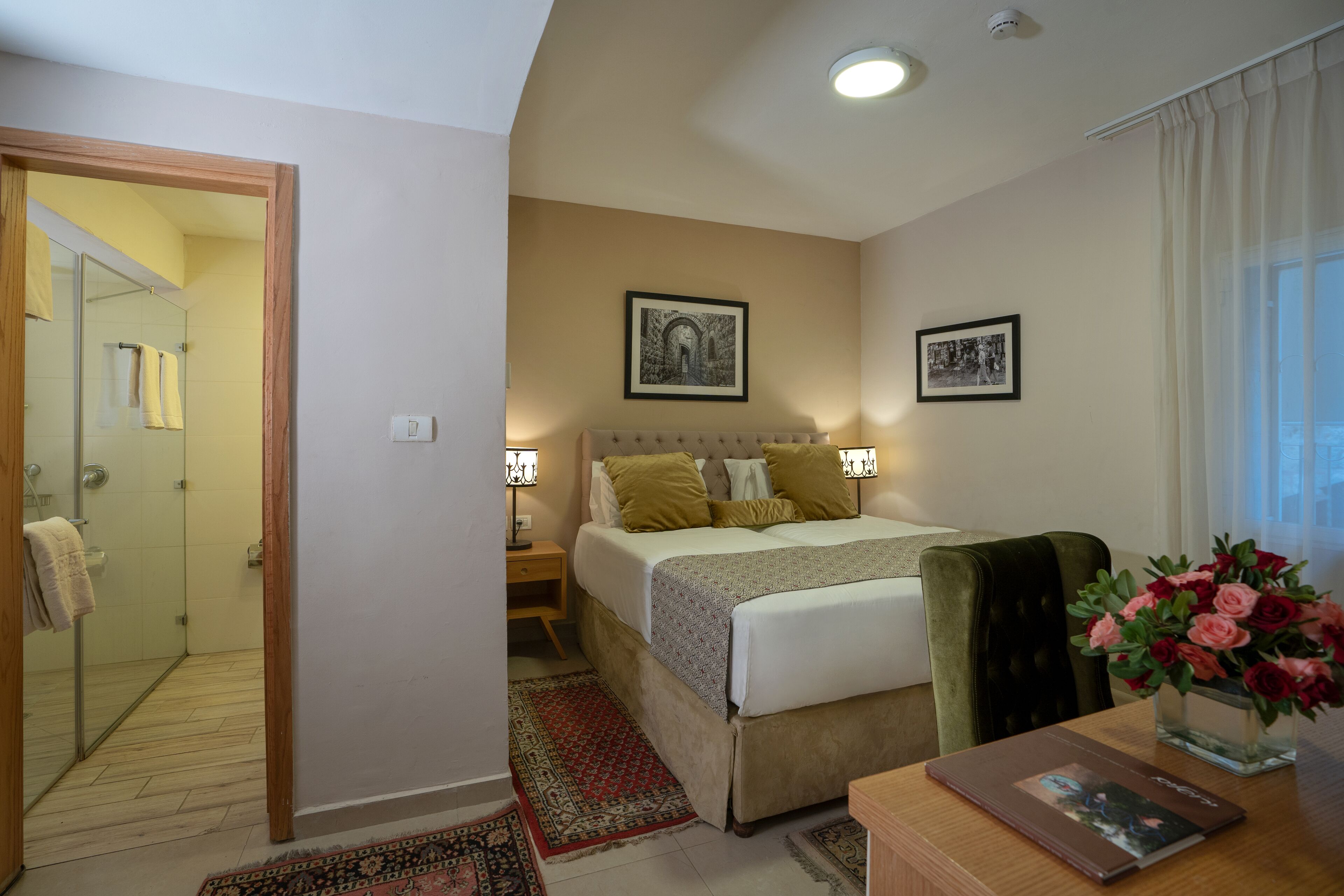 A hotel room with a bed, a nightstand, a desk, a chair, a desk lamp, a vase with flowers, a framed picture on the wall, and a shower area.