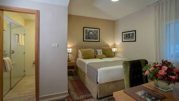 A hotel room with a bed, a nightstand, a desk, a chair, a desk lamp, a vase with flowers, a framed picture on the wall, and a shower area.