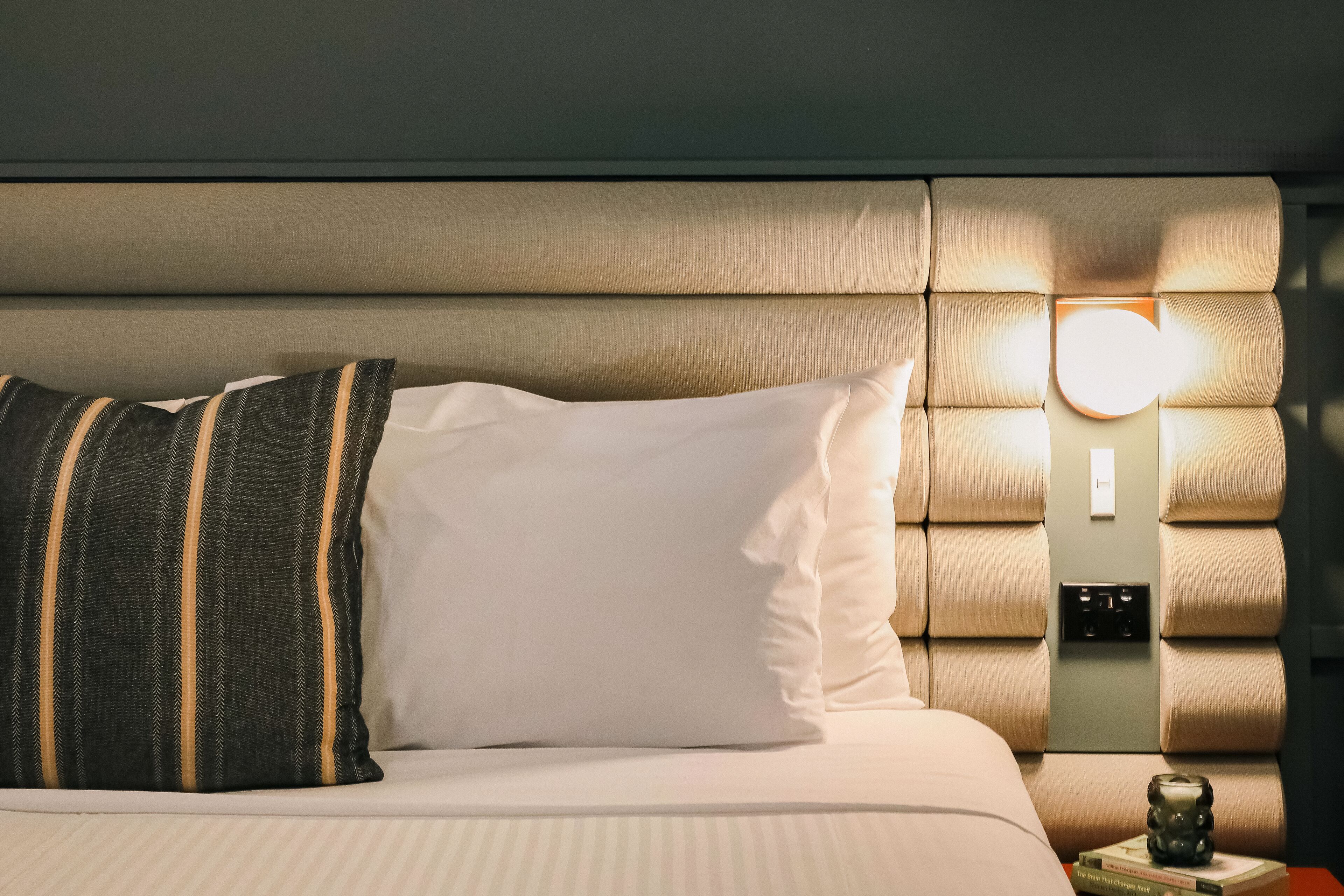 Ein ordentlich bezogenes Bett mit gestreiften und schlichten Kissen, ein Nachttisch mit Lampe und eine Wandleuchte.