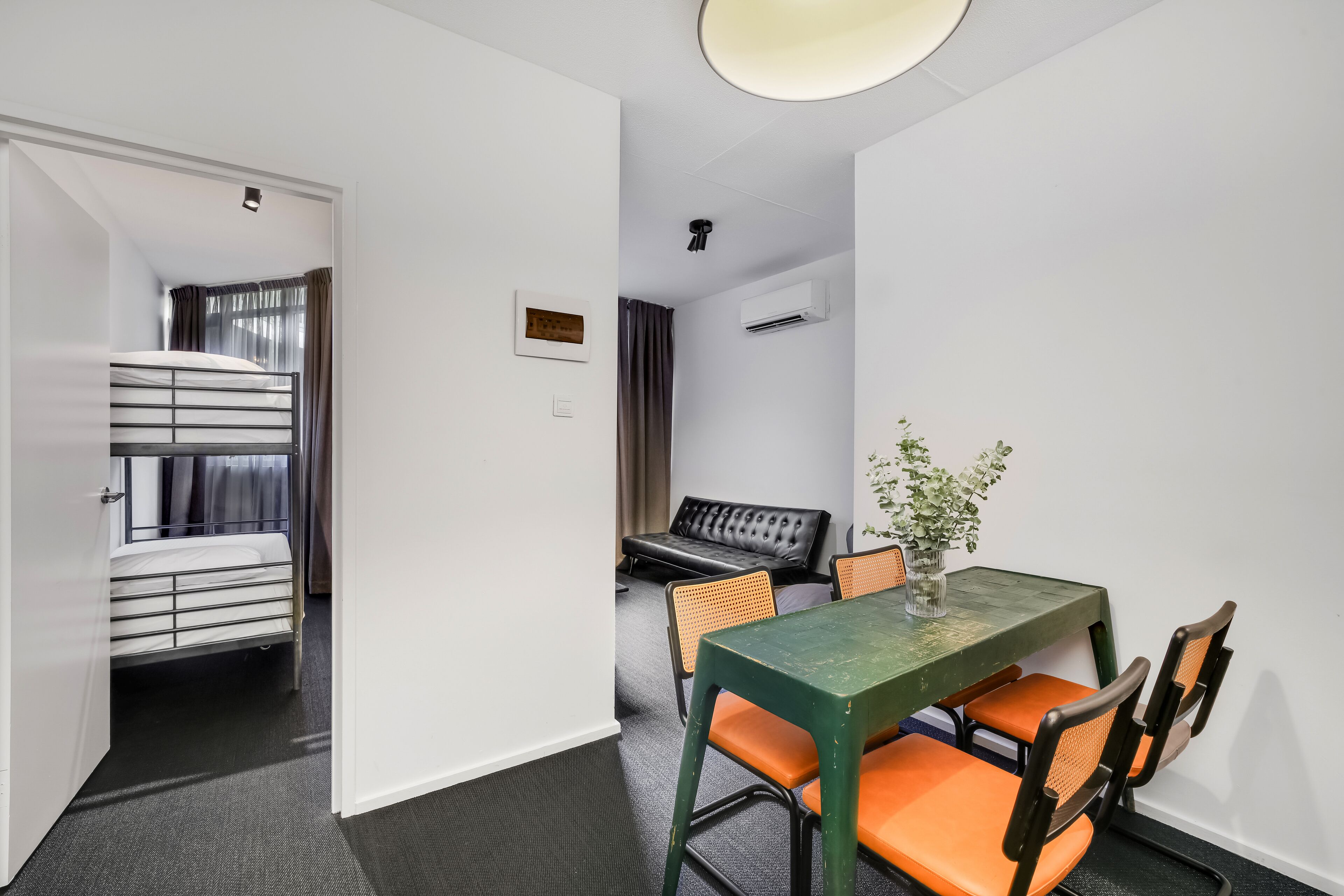 A compact hotel room with a bunk bed, a dining table with chairs, a sofa, and an air conditioning unit.