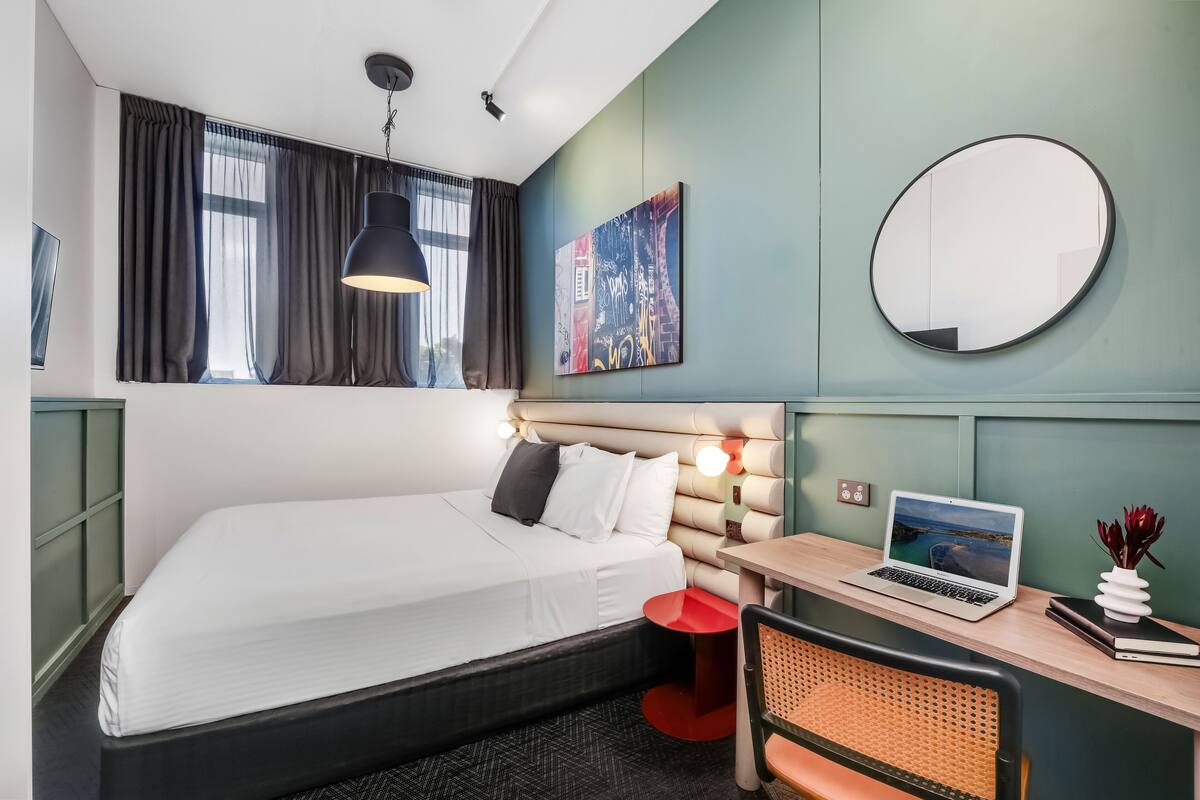 A modern hotel room with a bed, a desk with a laptop, a round mirror, and a painting on the wall.
