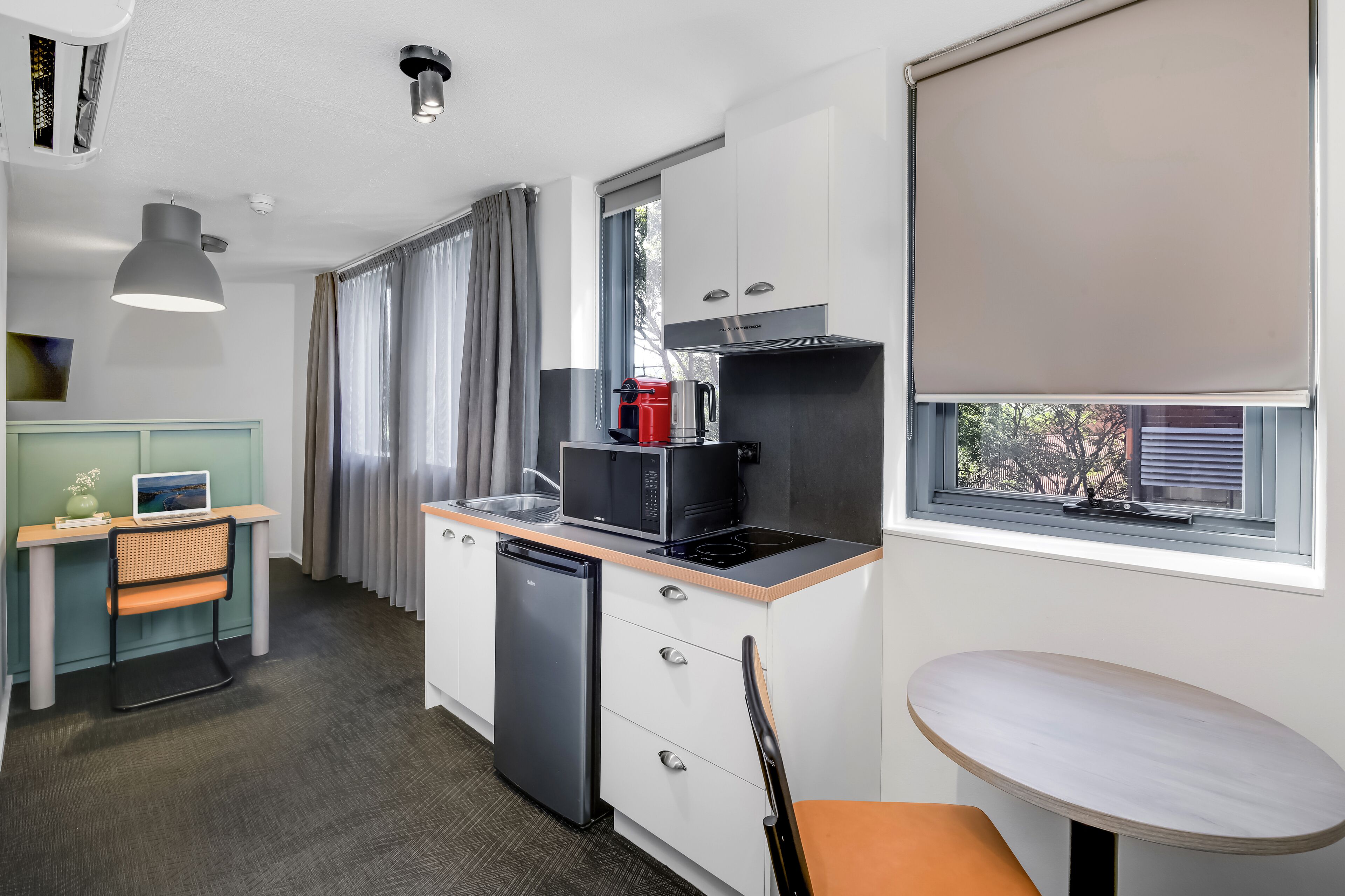 A compact kitchen with a refrigerator, microwave, and coffee maker. There is a dining table and a chair, and a window with a view of the outside.