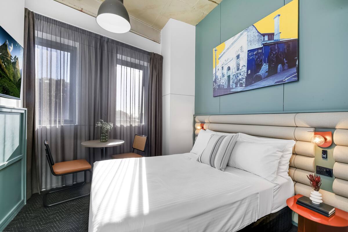 A hotel room with a large bed, a small table with a chair, a window with curtains, and a wall-mounted artwork.