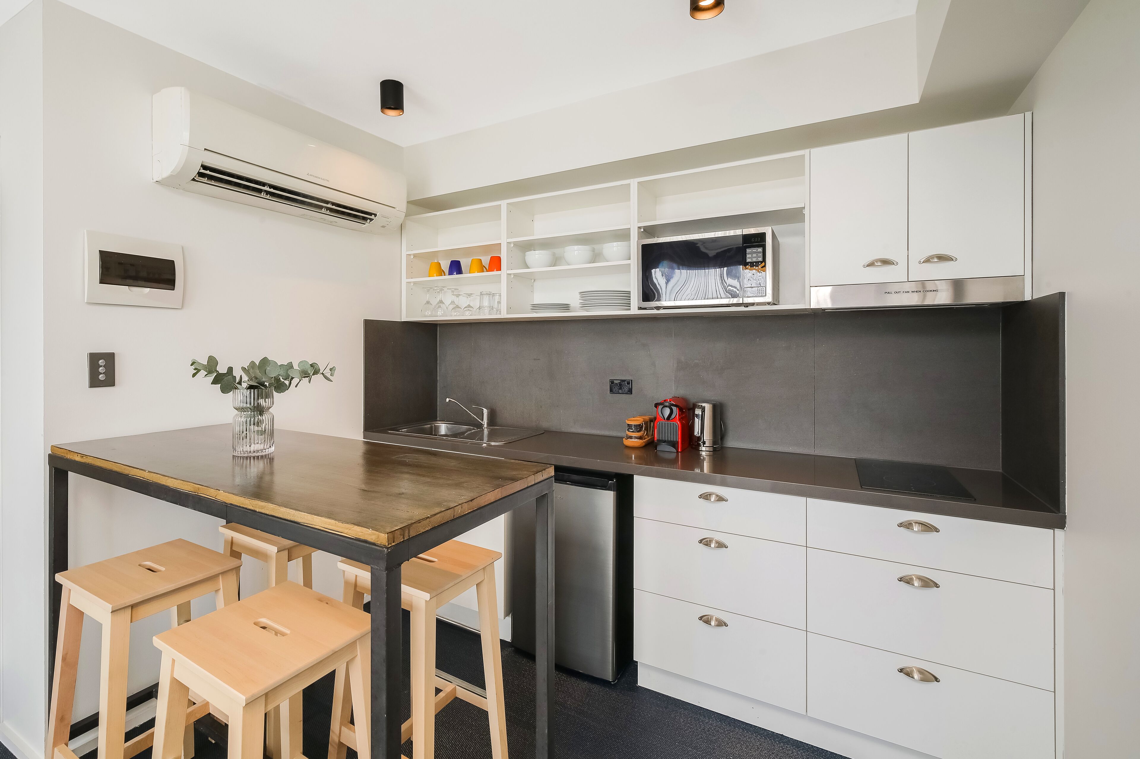 Cocina compacta con armarios blancos, microondas integrado y refrigerador pequeño. Hay una mesa de madera con dos taburetes y un florero con hojas verdes.