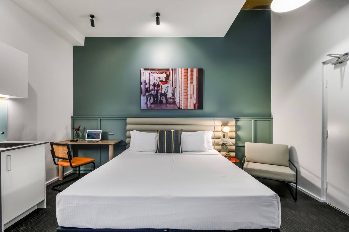 A hotel room with a large bed, a desk with a laptop, a chair, a sofa, and a wall-mounted artwork.