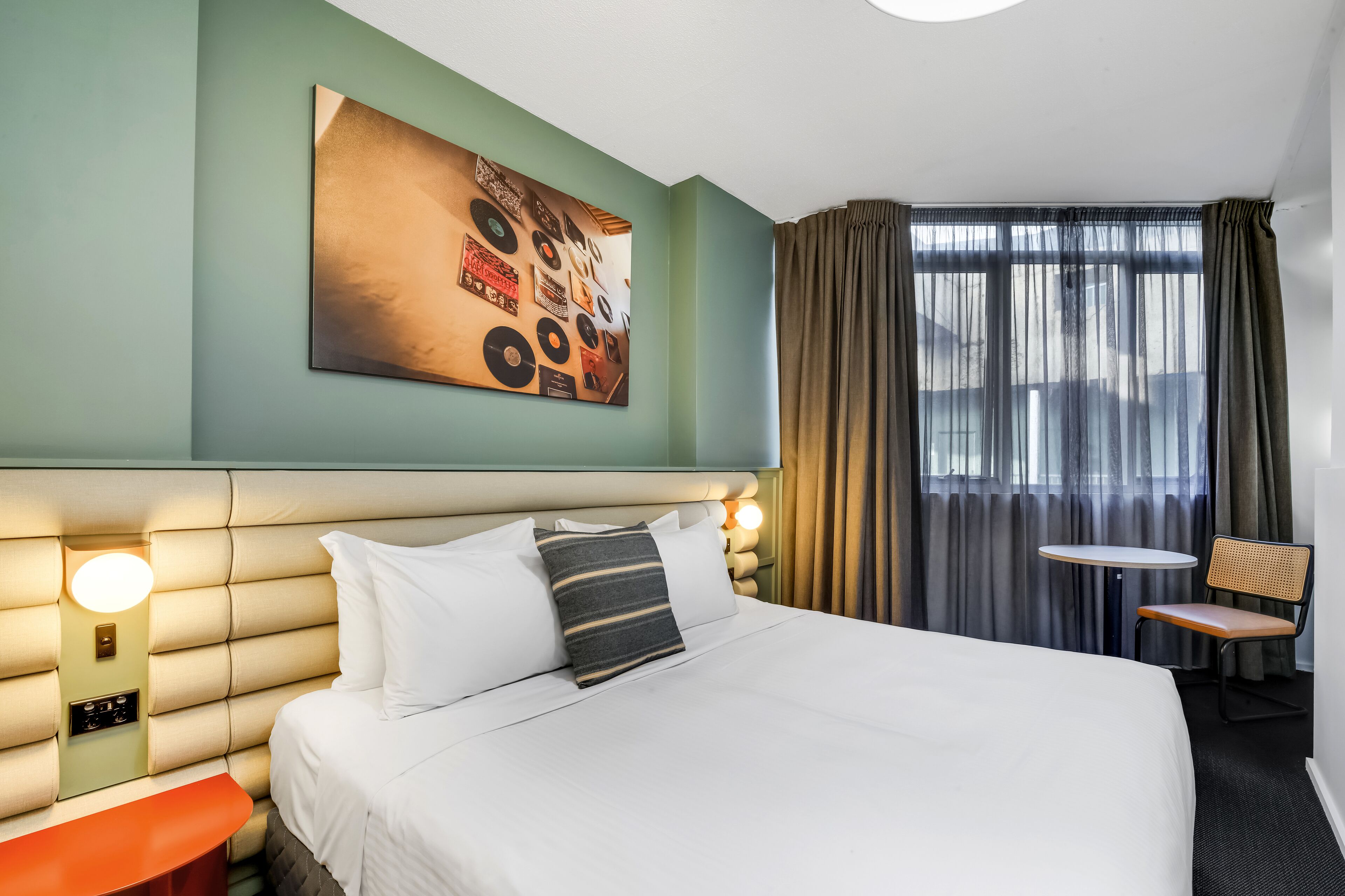 Ein Hotelzimmer mit einem großen Bett, einer Kopfteilbeleuchtung, einem Wandbild, einem kleinen Tisch mit Stuhl und einem Fenster mit Vorhängen.