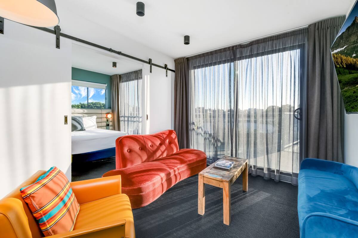 A modern hotel room with a bed, red and blue sofas, a small wooden table, and large windows with curtains.