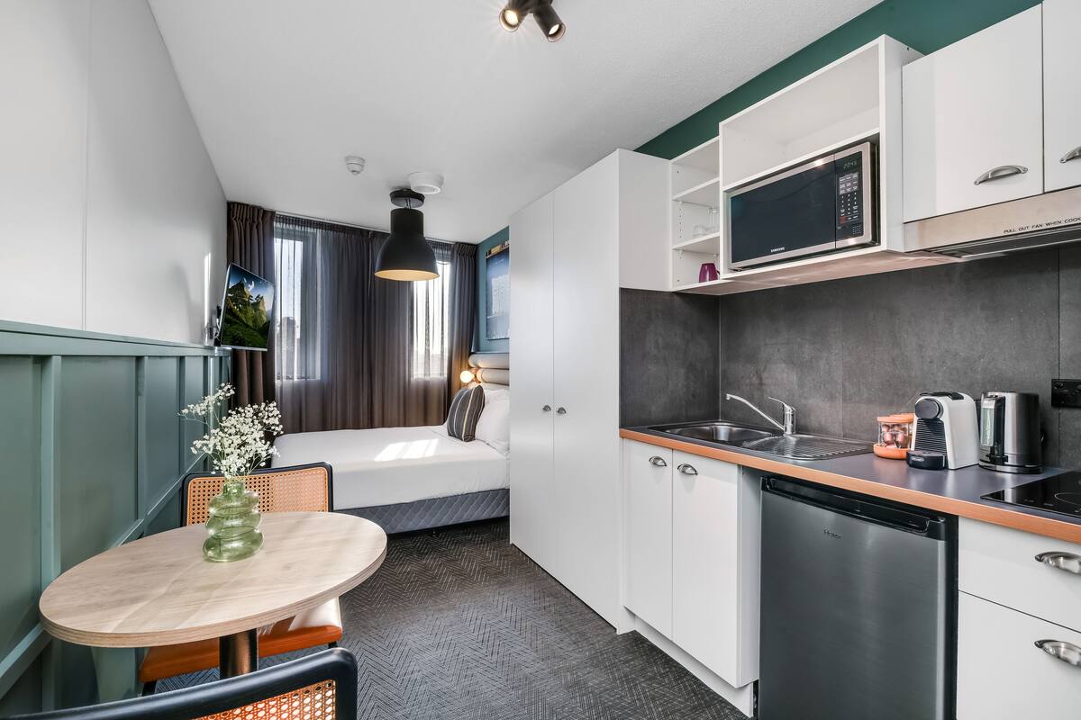 A compact hotel room with a kitchenette, a bed with a view, a dining area, and a bathroom.