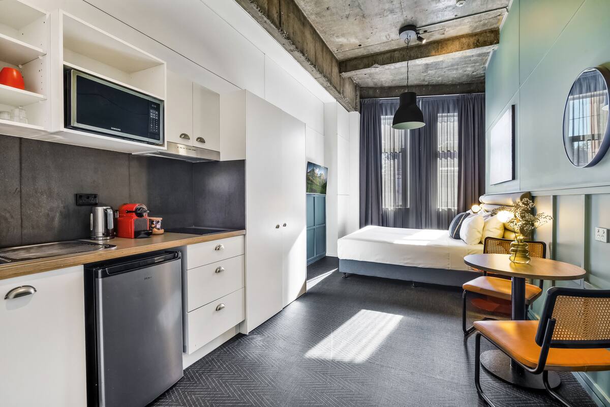 A compact hotel room with a kitchenette, a bed, a small dining area, and a view of the city.