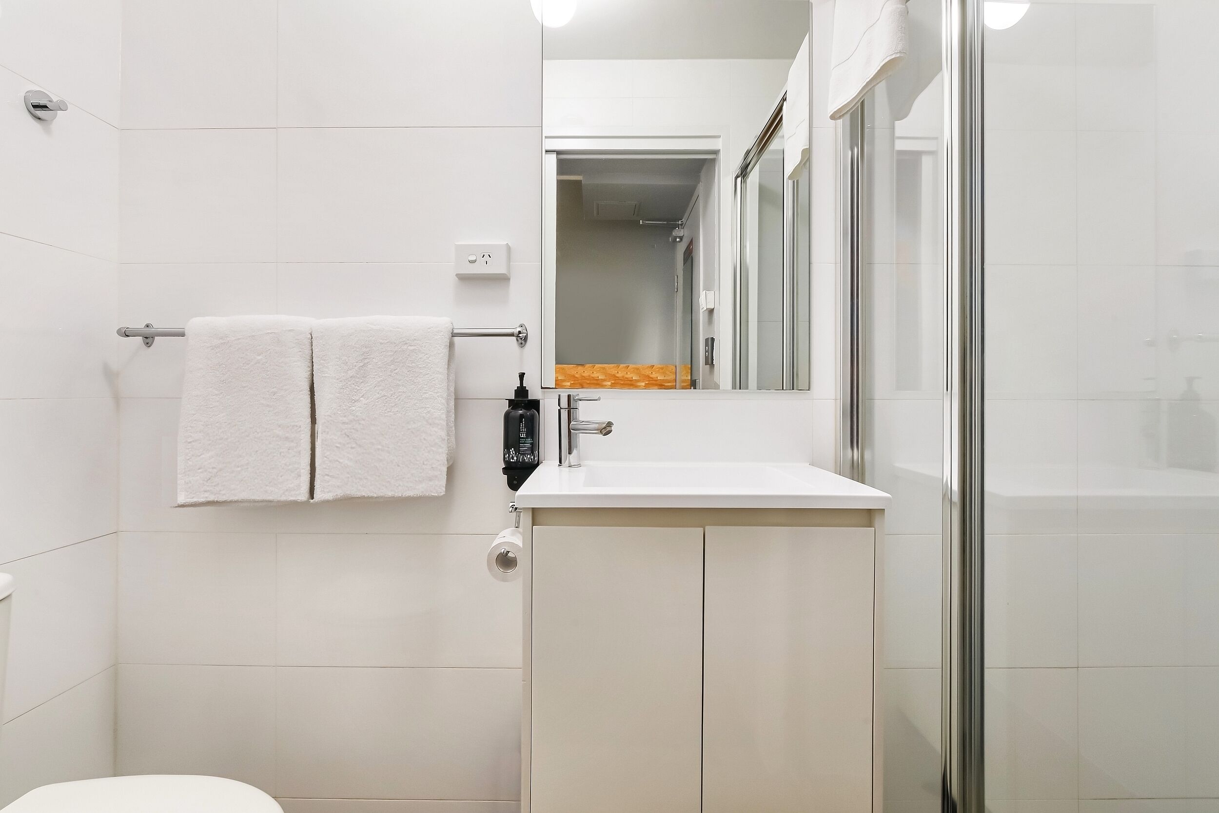 A modern bathroom with white tiles, a glass shower enclosure, a white sink with a mirror, and a towel rack.