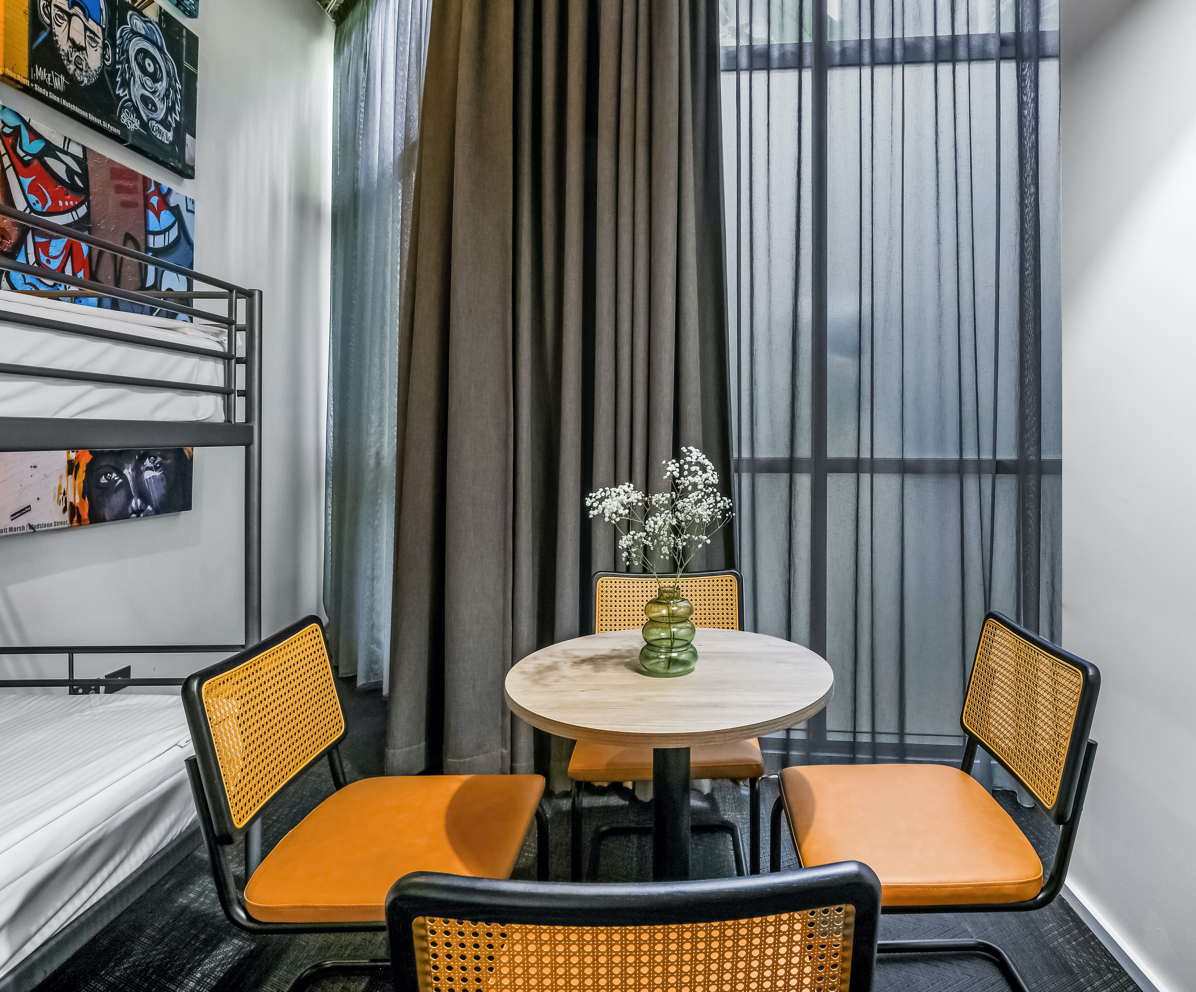 Een kamer met een bed, een ronde tafel met twee stoelen, een vaas met bloemen en een raam met gordijnen.