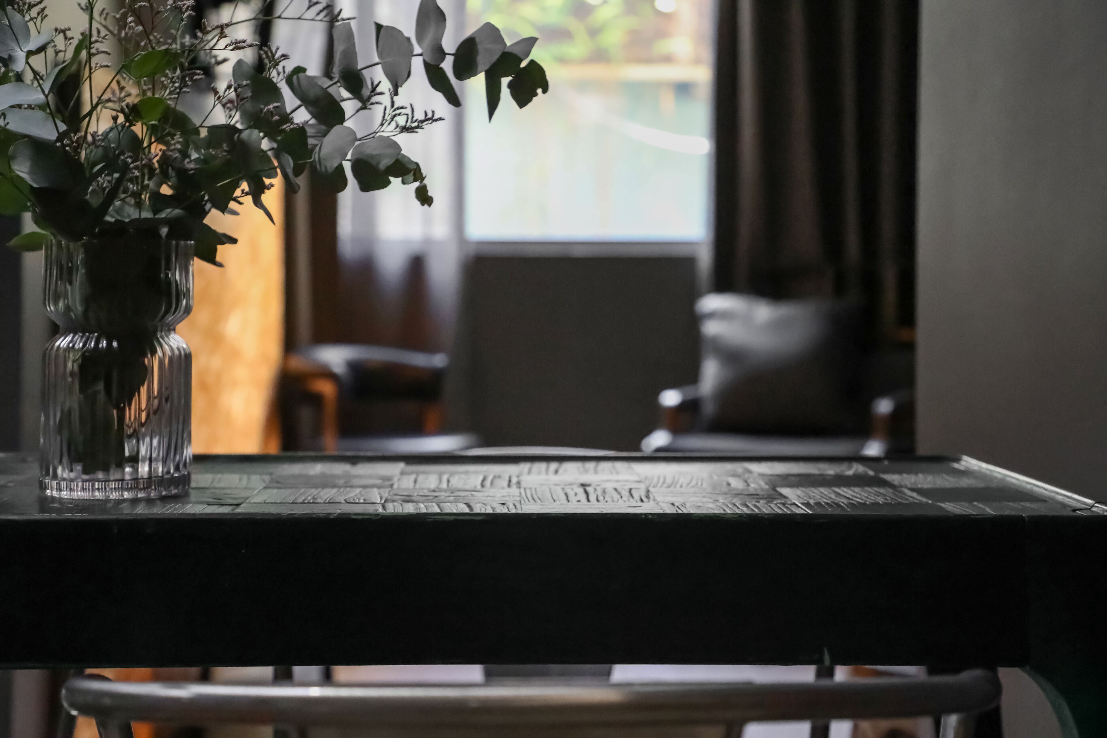 A clear view of a table with a vase of green plants, a wooden table surface, and a window in the background.