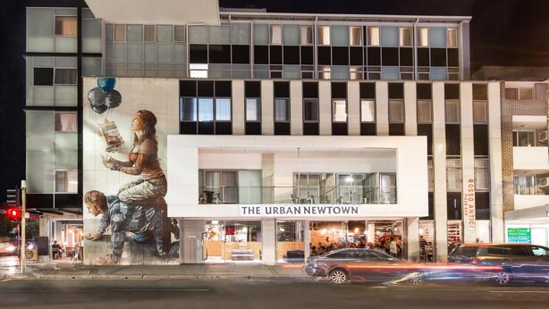 Un bâtiment moderne, orné d’une grande fresque sur sa façade, avec une enseigne indiquant « The Urban Newtown », et une entrée bien visible où l’on peut voir des gens entrer.