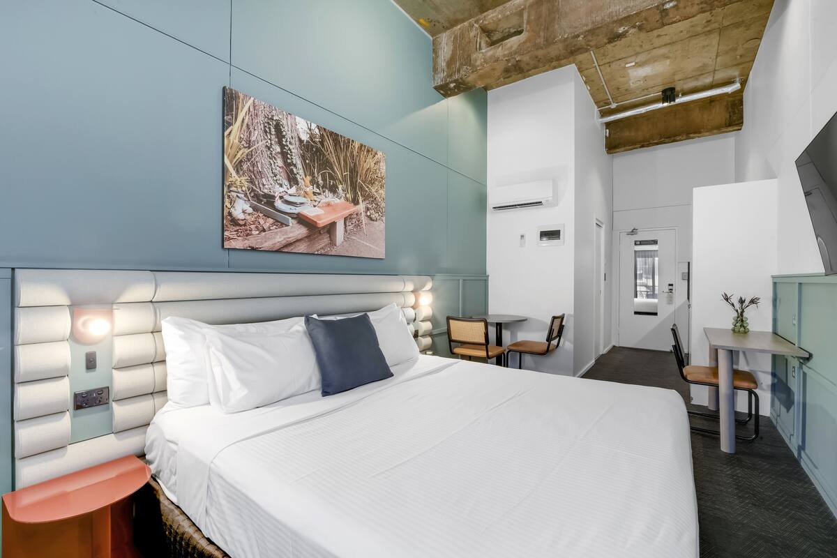 A modern hotel room with a large bed, a headboard with built-in lighting, a small table with chairs, and a painting on the wall.