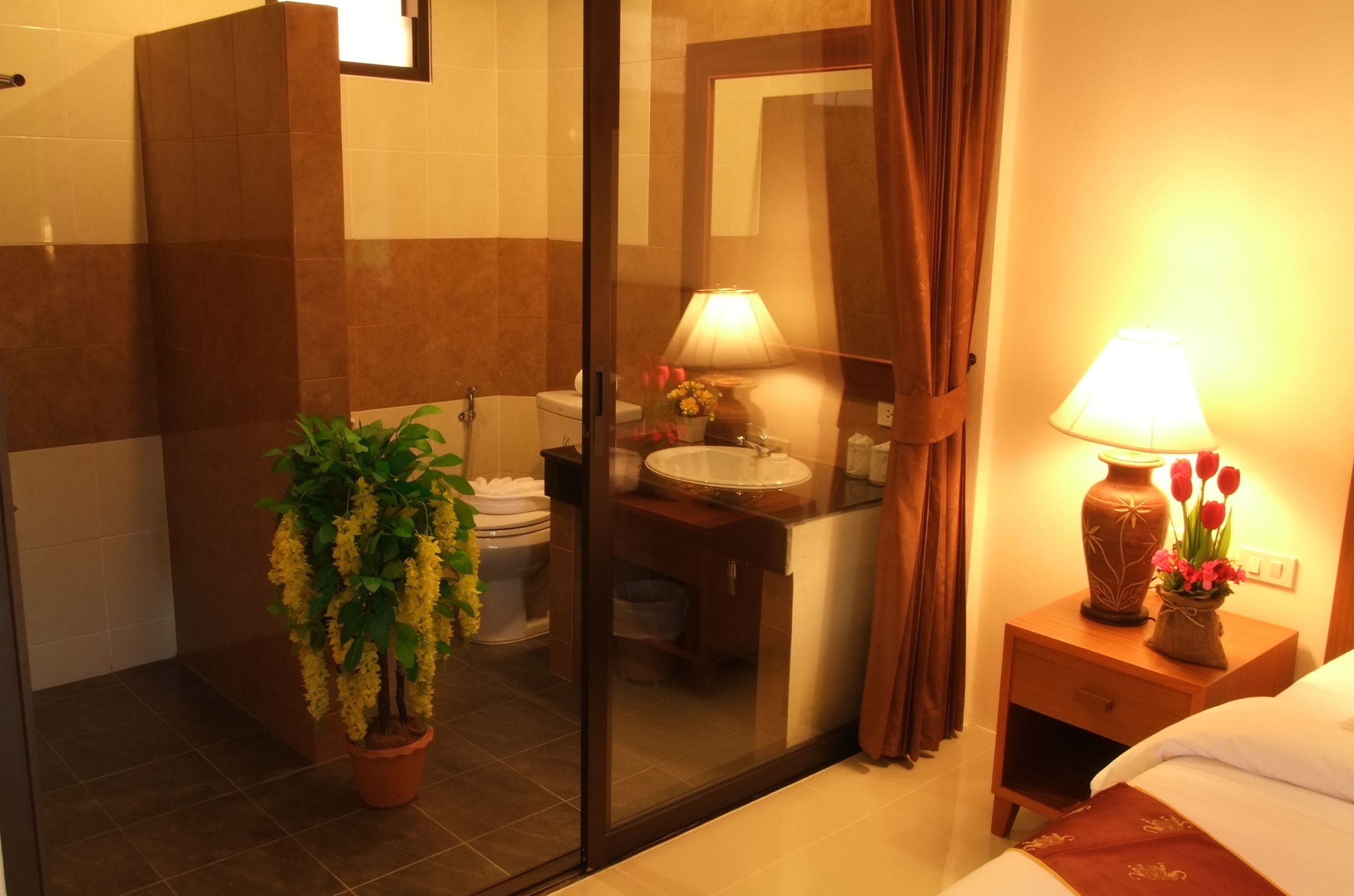 A hotel bathroom with a glass shower enclosure, a sink with a mirror, and a bedside table with a lamp and flowers.