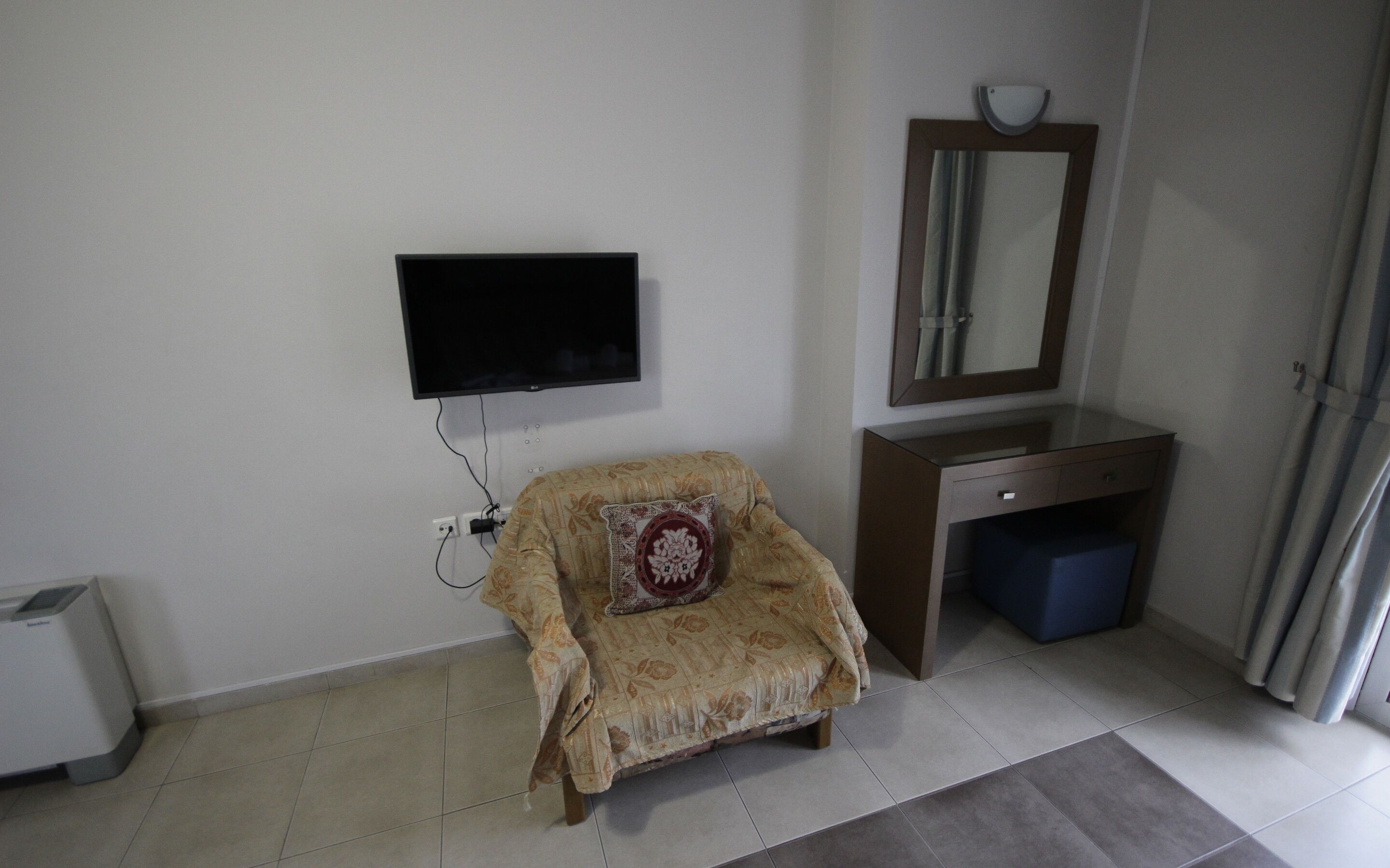 A room with a television mounted on the wall, a small wooden chair with a cushion, a desk with a mirror, and a blue trash bin.