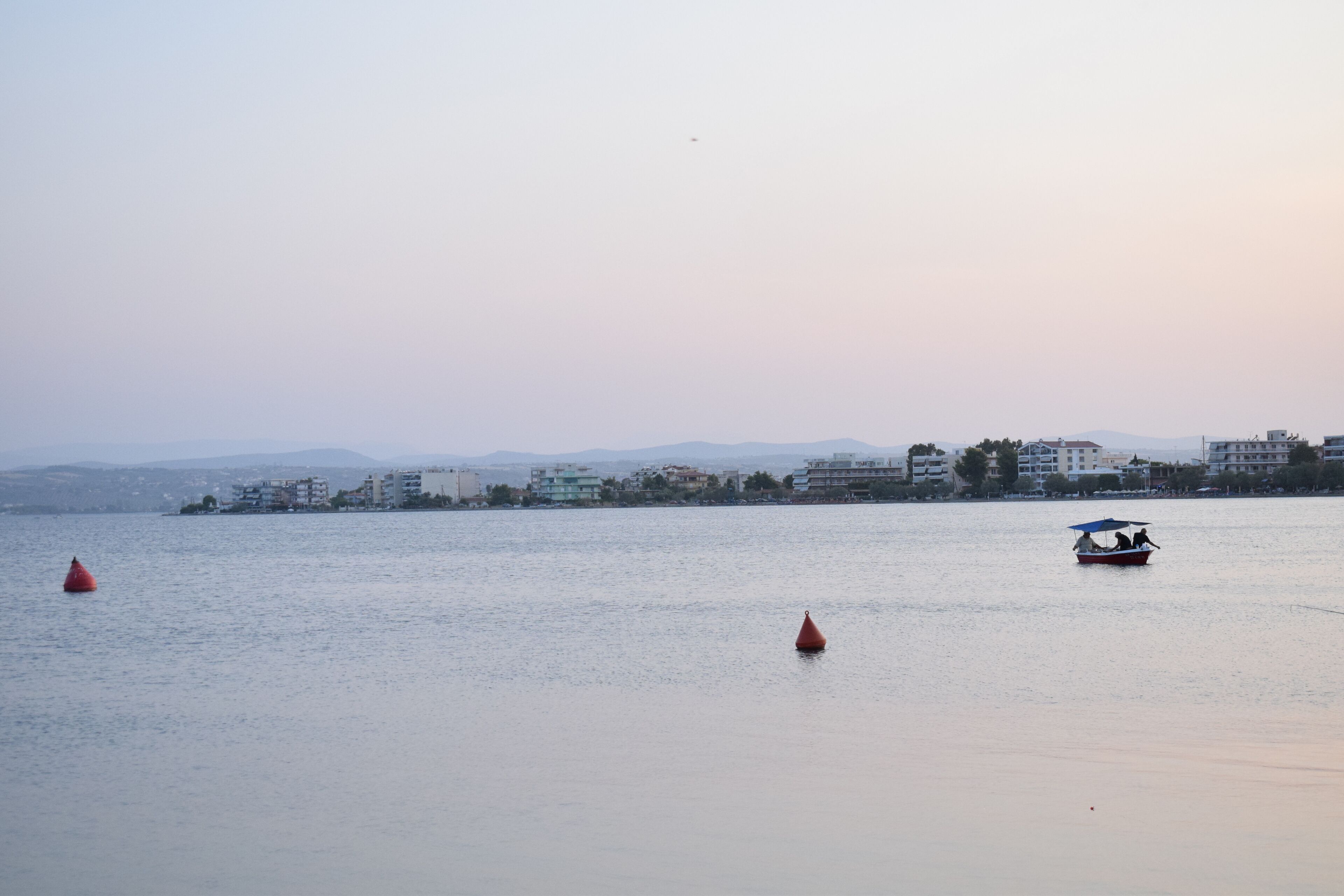 A calm body of water with a boat carrying passengers, buoys, and buildings in the background.