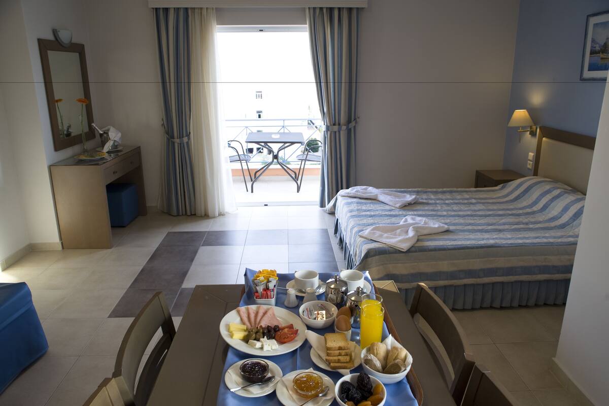 Habitación de hotel con una mesa de comedor dispuesta para el desayuno, una cama con ropa de cama a rayas, una mesita de noche con lámpara y vistas al balcón exterior.