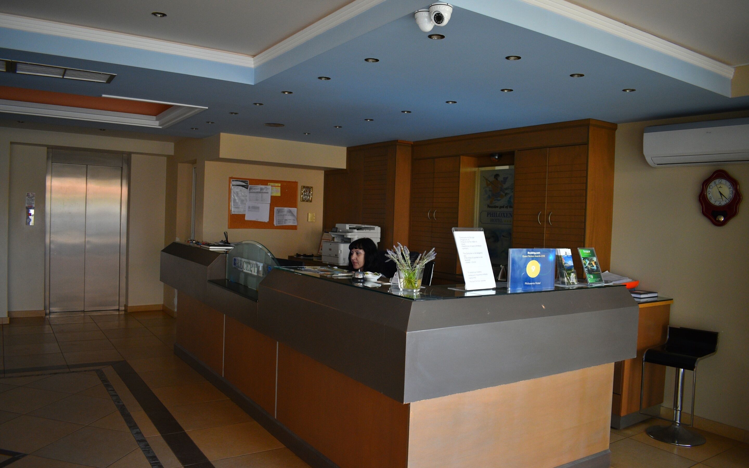 A hotel reception area with a reception desk, a bulletin board, a clock, and an elevator.