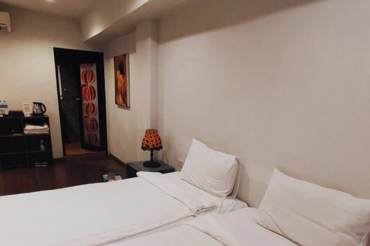 A hotel room with a large bed, a nightstand with a lamp, a desk with a chair, and a door leading to another room.