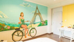 Una habitación con un mural de la Torre Eiffel y una mujer en bicicleta, una cama, una mesita con mantel blanco y una puerta.