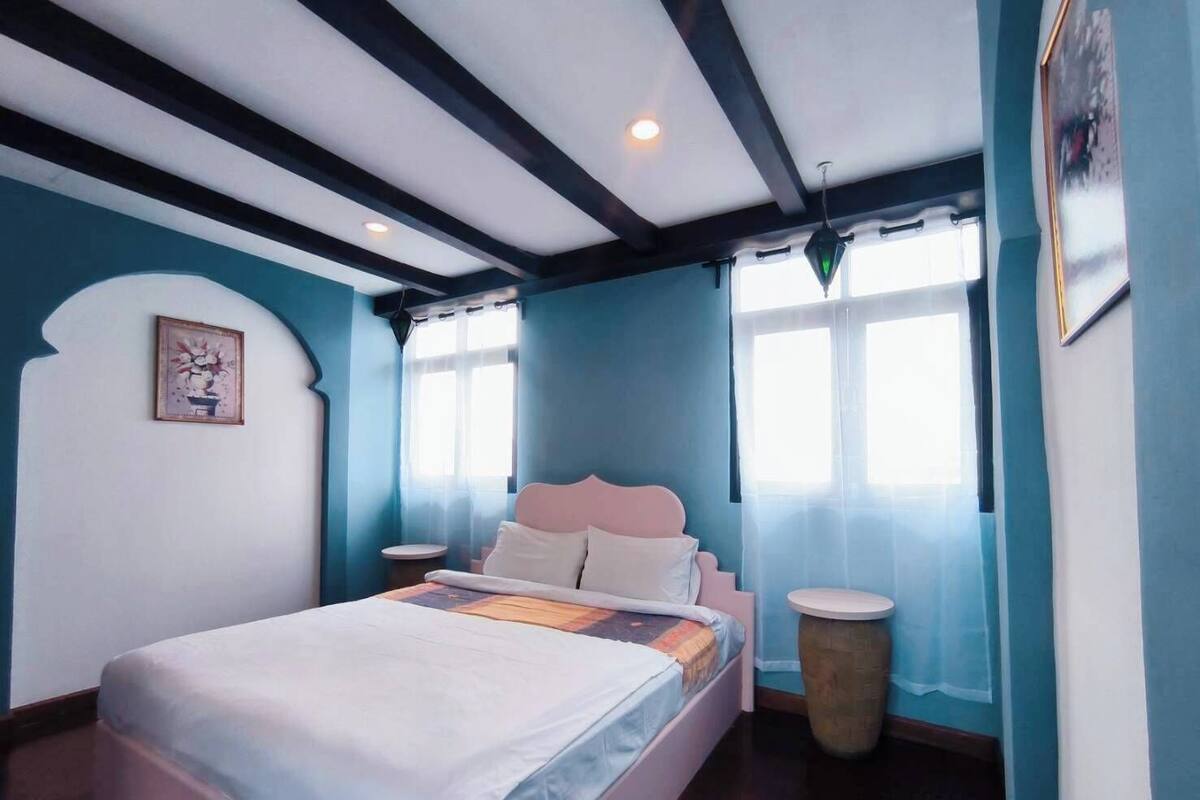 A bedroom with a bed, a round nightstand, a wall-mounted picture, and a window with curtains.