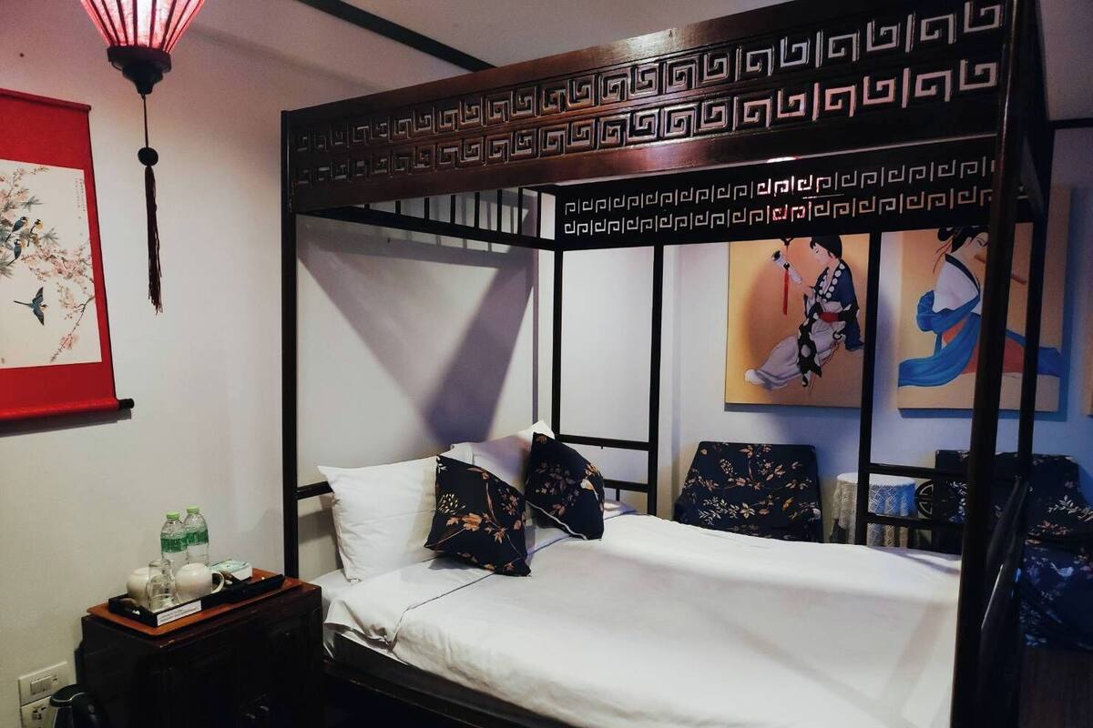 A four-poster bed with white linens and floral pillows, a wooden headboard with a geometric pattern, a red framed artwork on the wall, and a small bedside table with a kettle and bottles.