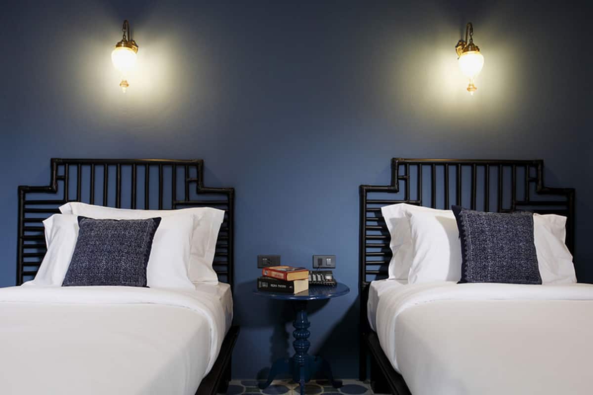 Two beds with white linens and dark pillows, a small table with books and a lamp, and wall-mounted lights.