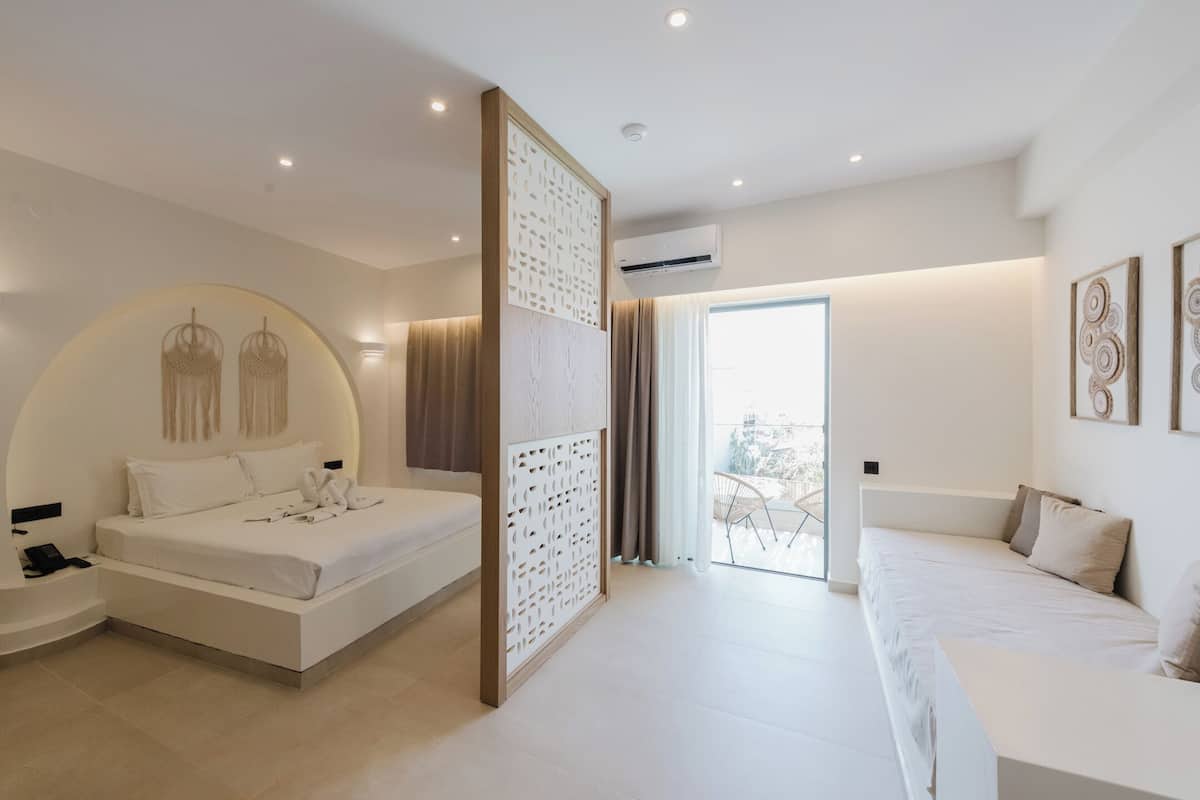 A modern hotel room with a large bed, a smaller sofa, a decorative wall panel, and a balcony with a view.