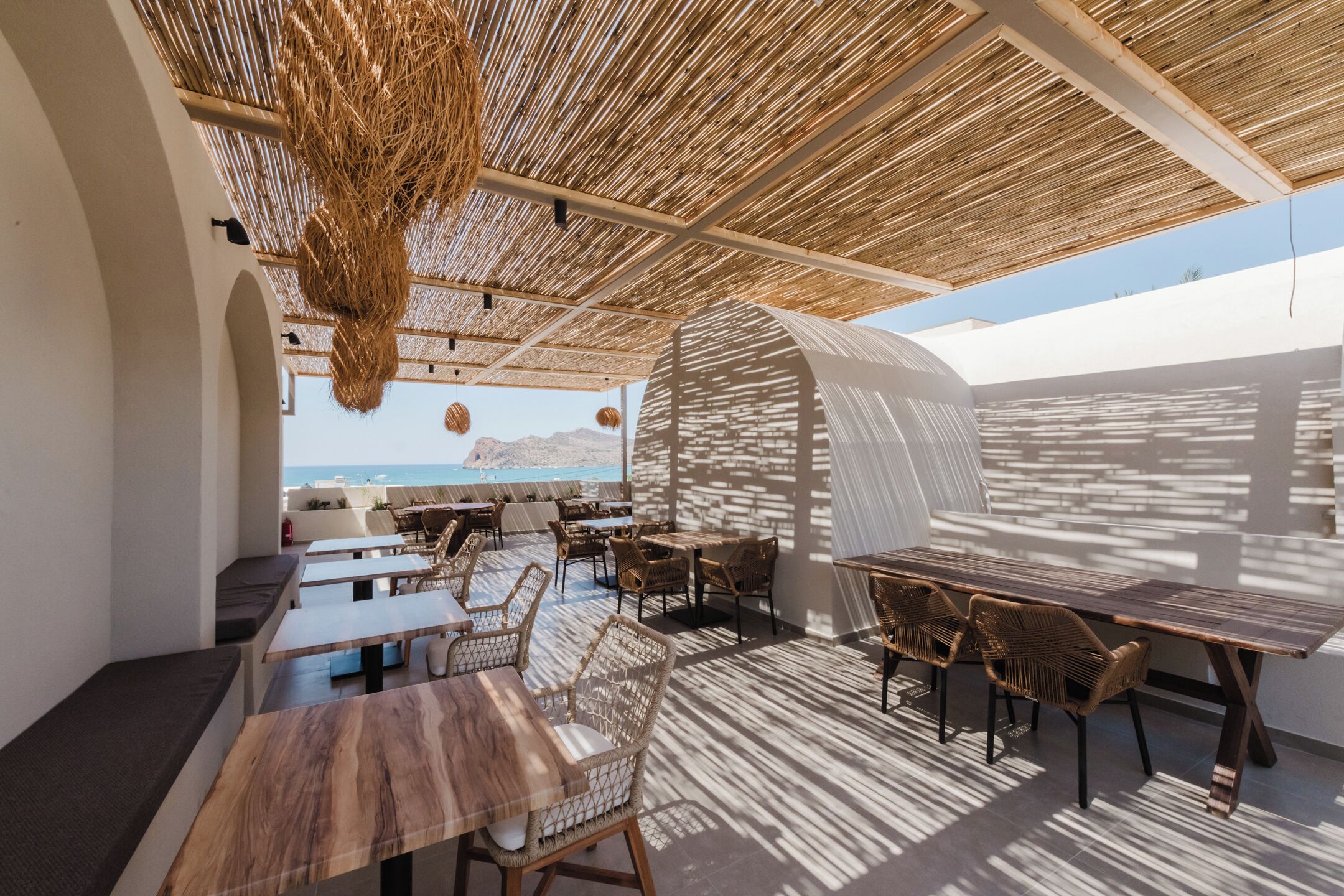 An outdoor dining area with wooden tables and wicker chairs, overlooking a beach.