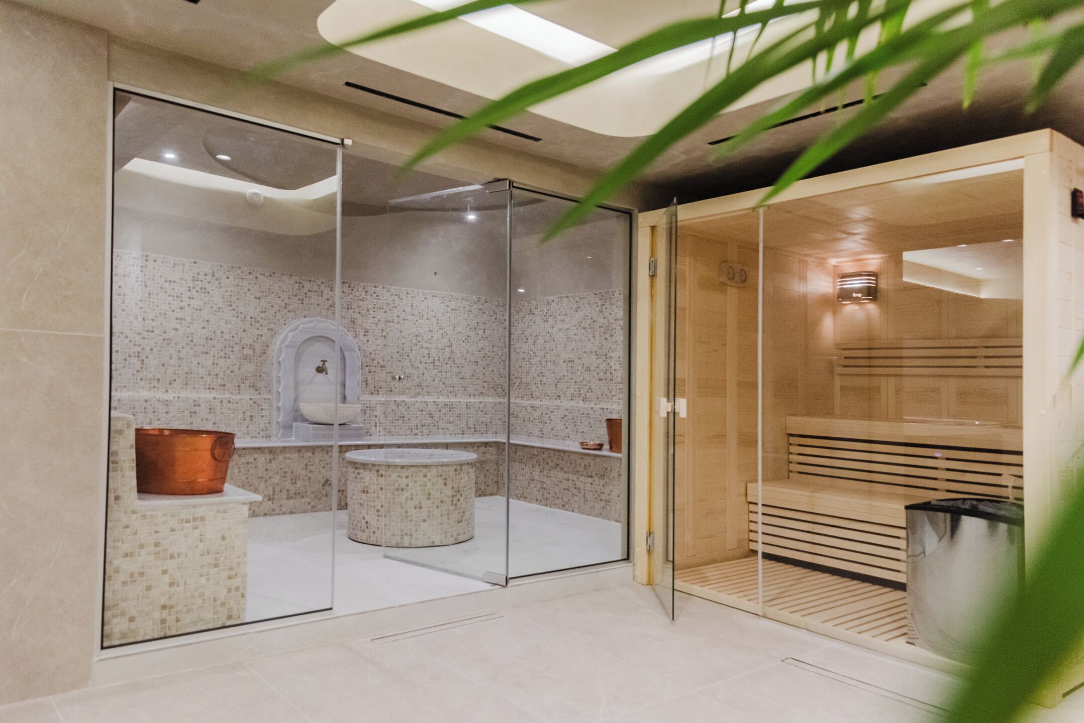 Un moderno cuarto de spa con una sauna de paredes de vidrio, un banco de madera y un lavabo de cobre.