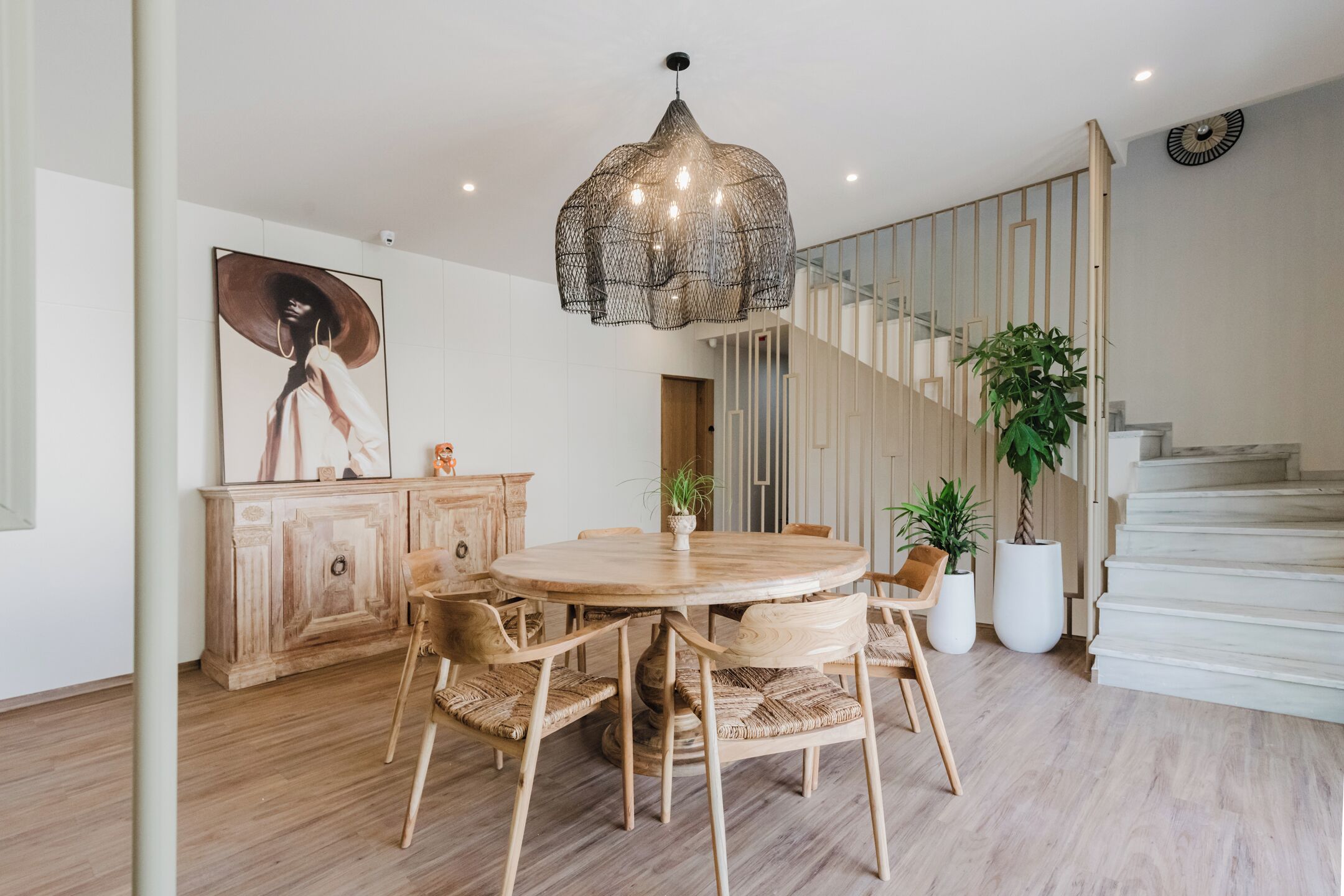 A dining area with a round wooden table, wicker chairs, a hanging light fixture, a staircase, and potted plants.