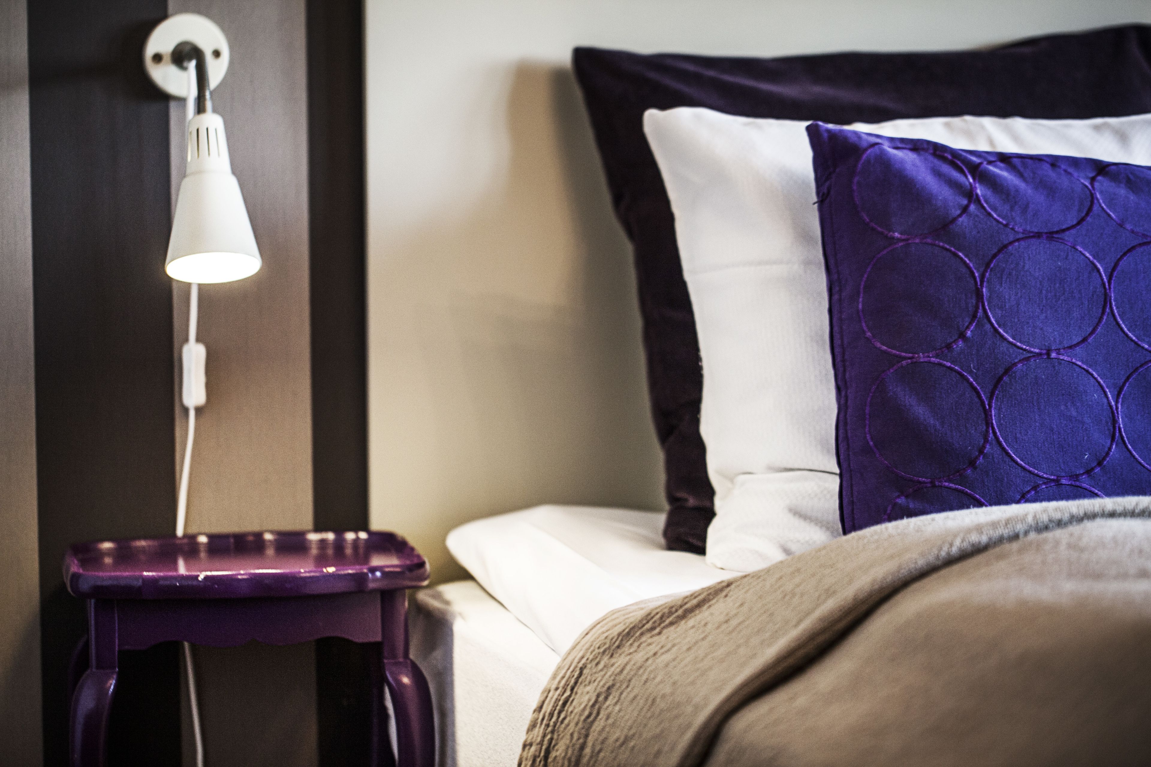 Ein Bett mit einem weißen Kopfteil, einem violetten Kissen und einem Nachttisch mit einer Lampe.