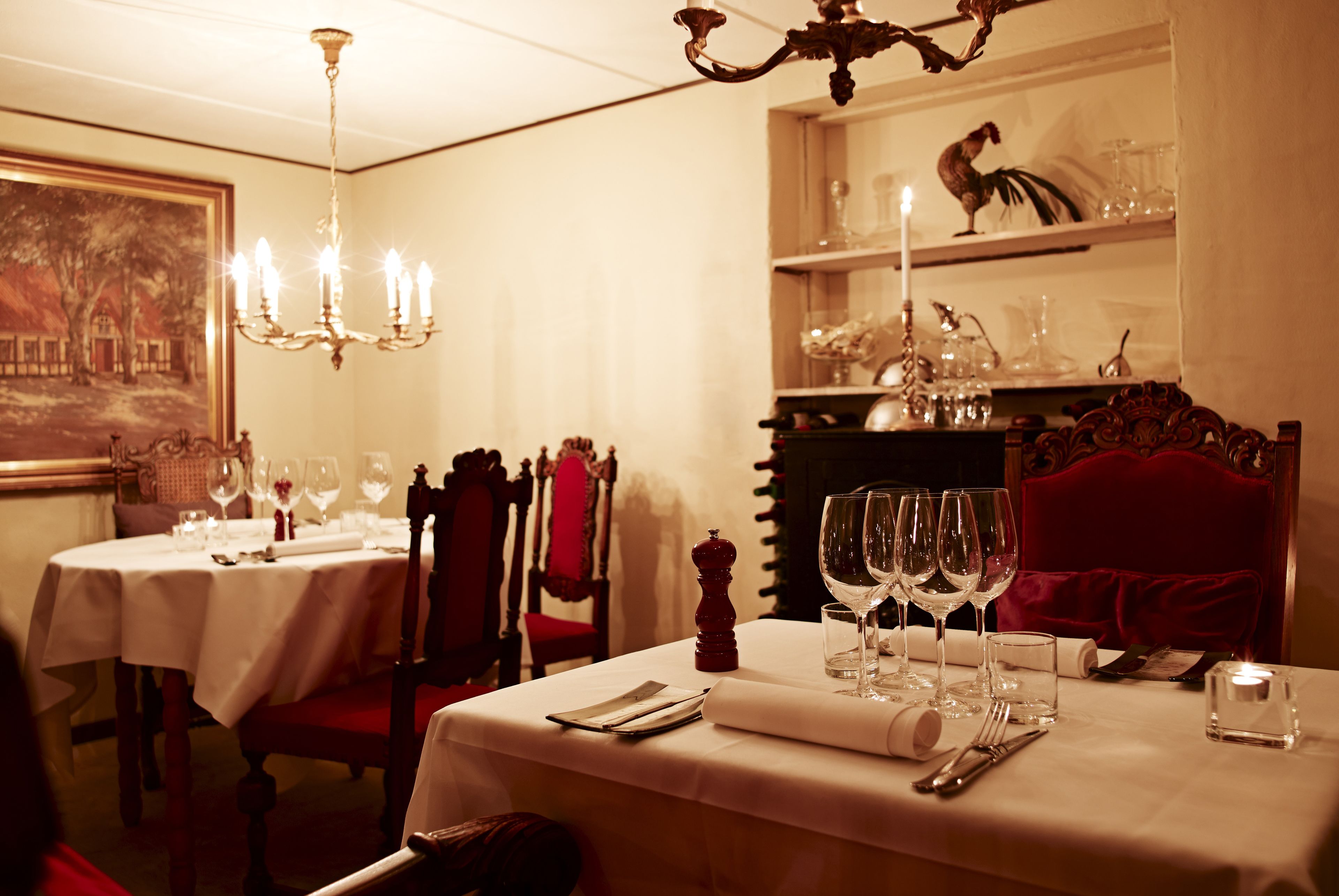 A dining area with a table set for two, a chandelier, and a painting on the wall.