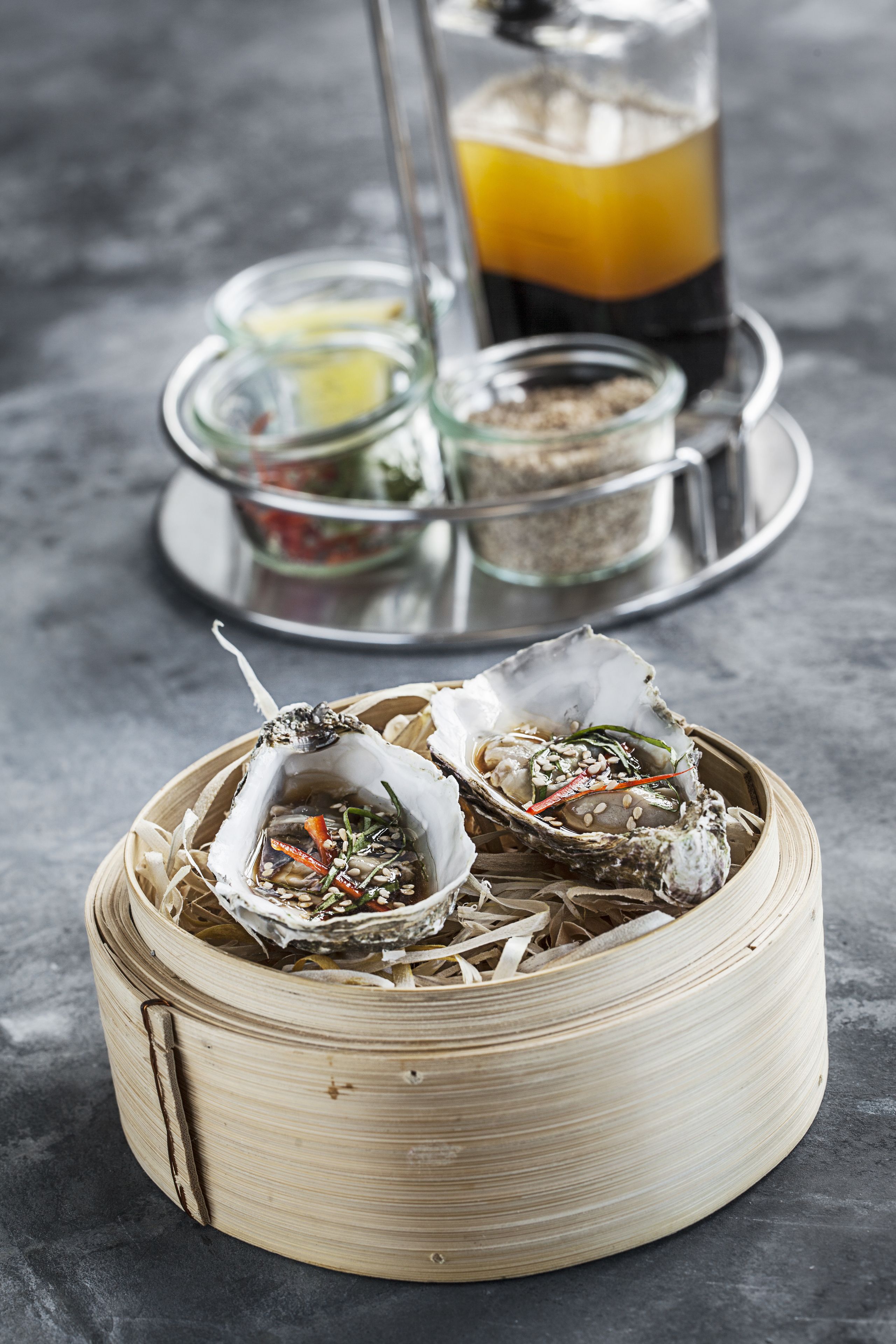A bamboo steamer with seafood, a glass of juice, and condiments on a metal tray.