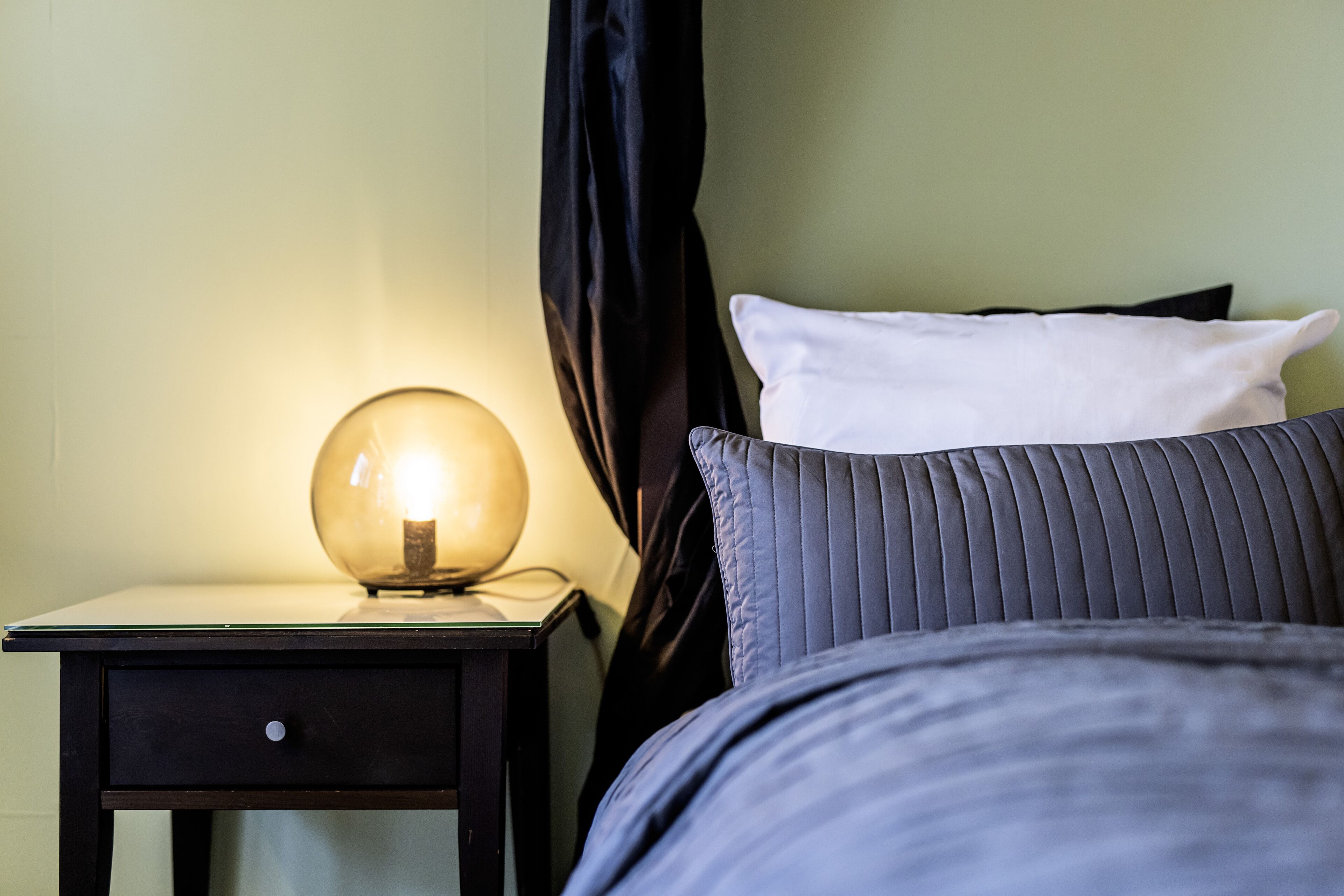 Una cama bien tendida con almohada gris y blanca, una lámpara redonda sobre una mesita de noche y una cortina negra parcialmente abierta.