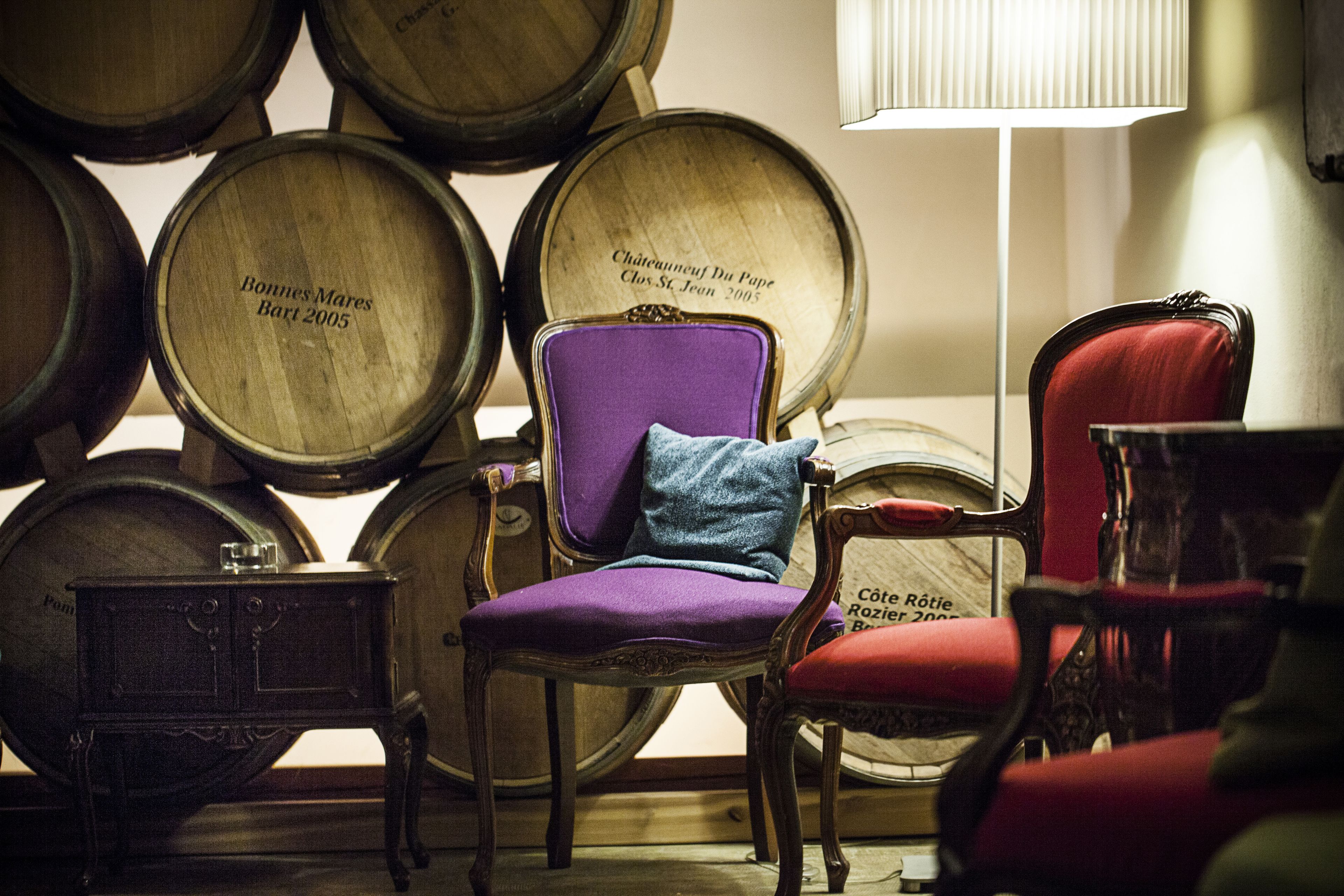 Una habitación con barriles de madera, un sillón morado con un cojín, un sillón rojo y una pequeña mesa.