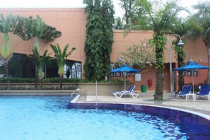 Une piscine entourée de verdure, agrémentée de chaises longues et de parasols, et bordée d’un bâtiment à la façade de couleur pêche.