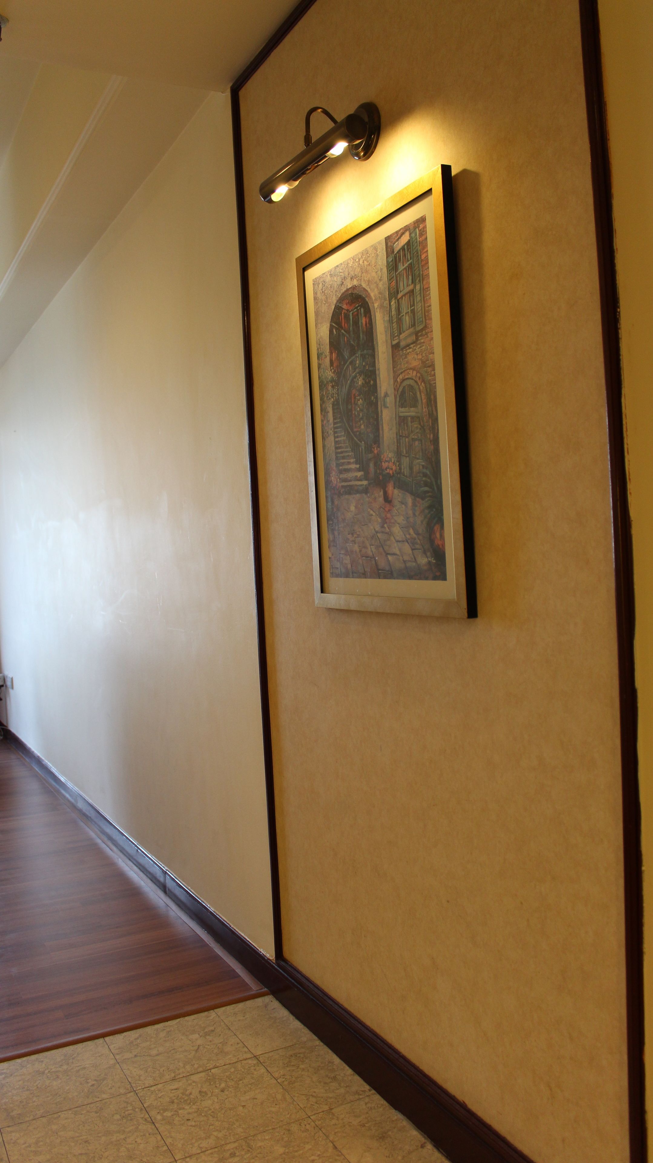 A hallway with a framed painting of a building, a wall-mounted light fixture, and a wooden floor.