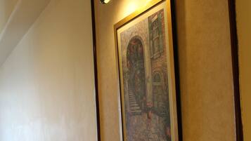 A hallway with a framed painting of a building, a wall-mounted light fixture, and a wooden floor.