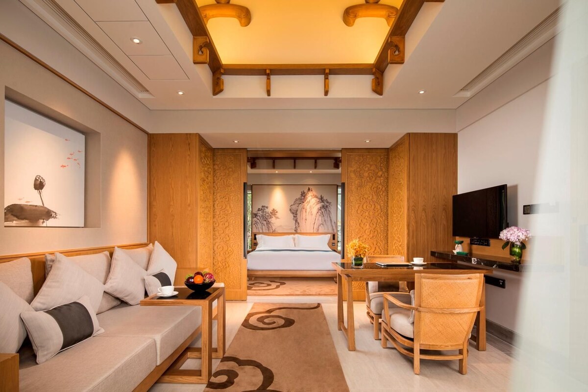 A modern hotel room with a sofa, dining table, chairs, a bed, and a television.