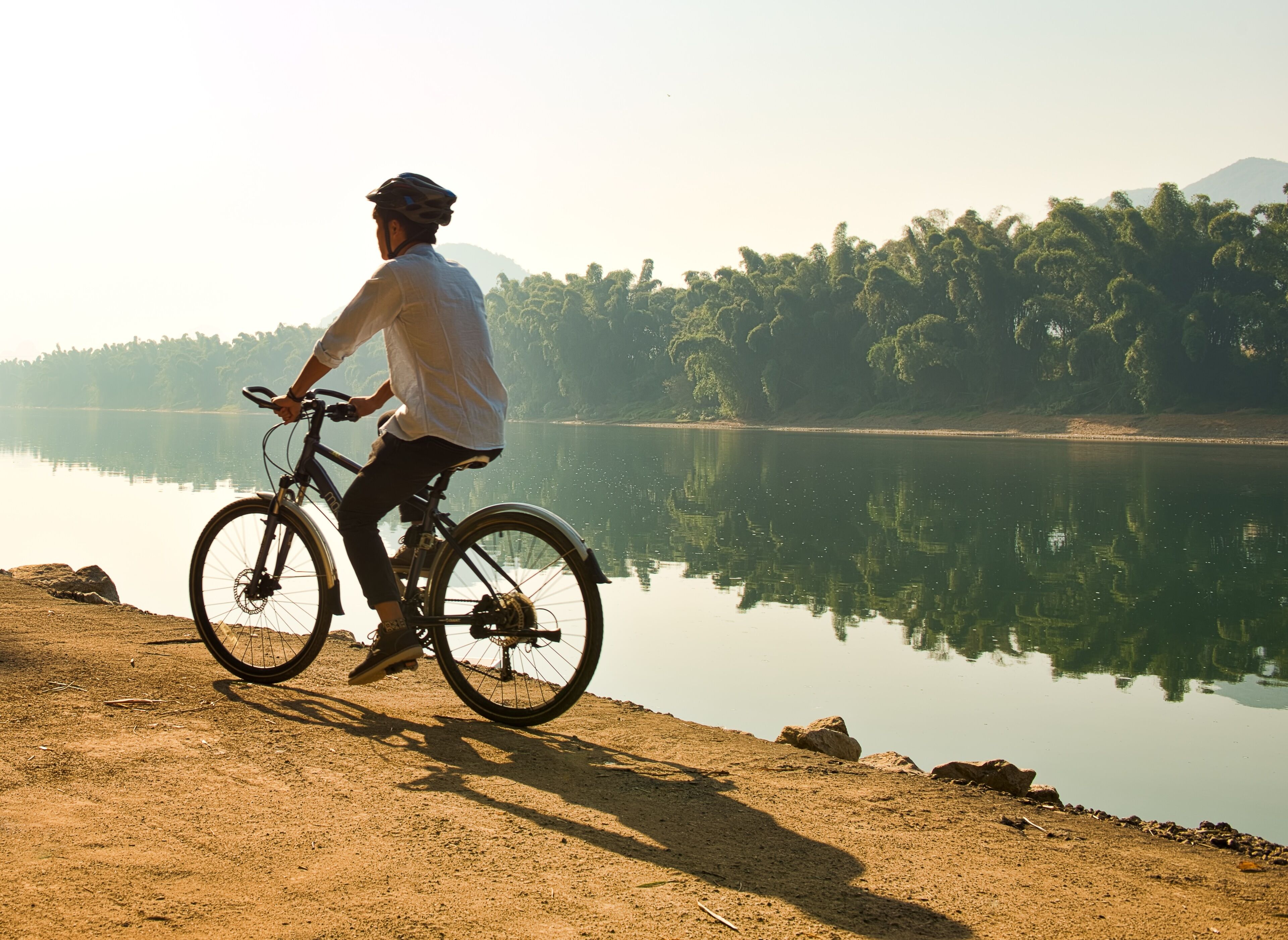 A person riding a bicycle on a dirt path by a calm lake with trees in the background.
