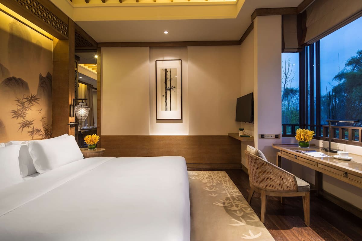 A hotel room with a large bed, a desk with a chair, a framed artwork on the wall, and a window with a view of trees.