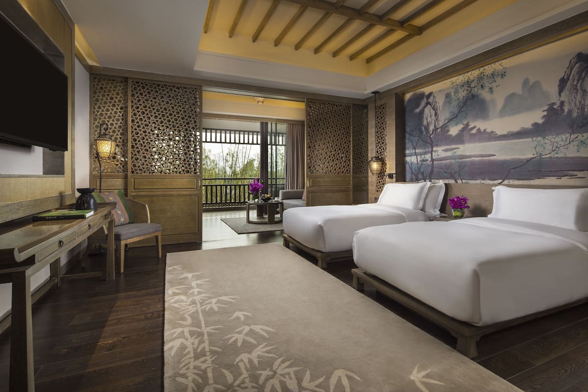 A hotel room with a wooden desk, two beds, a TV, and a large painting of a landscape.