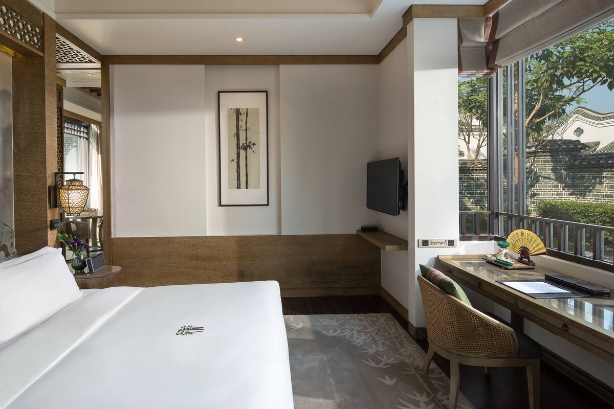 A hotel room with a bed, a desk with a chair, a TV, and a window with a view of a traditional building.