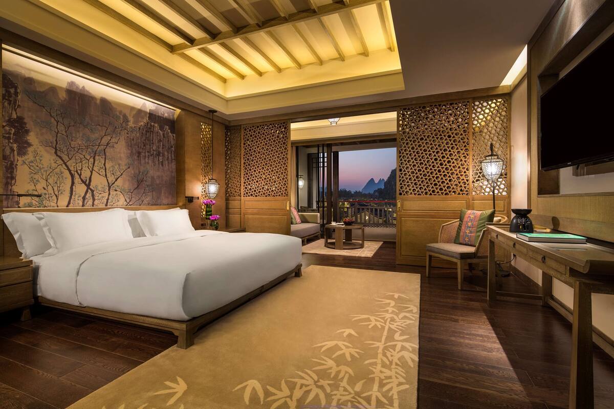 A hotel room with a large bed, a desk, a chair, a TV, and a view of the mountains.