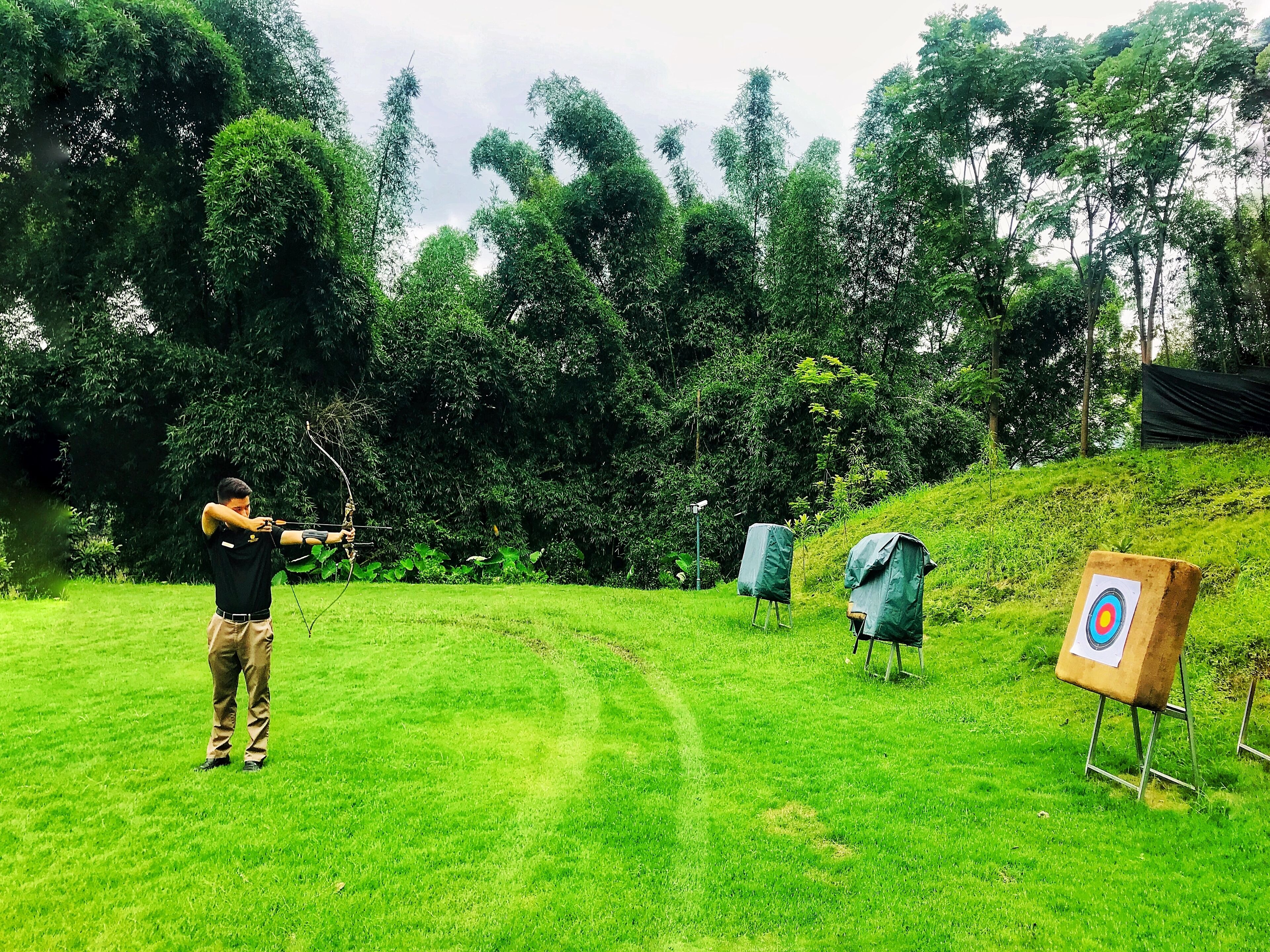 A person practicing archery in a grassy outdoor area with trees in the background.